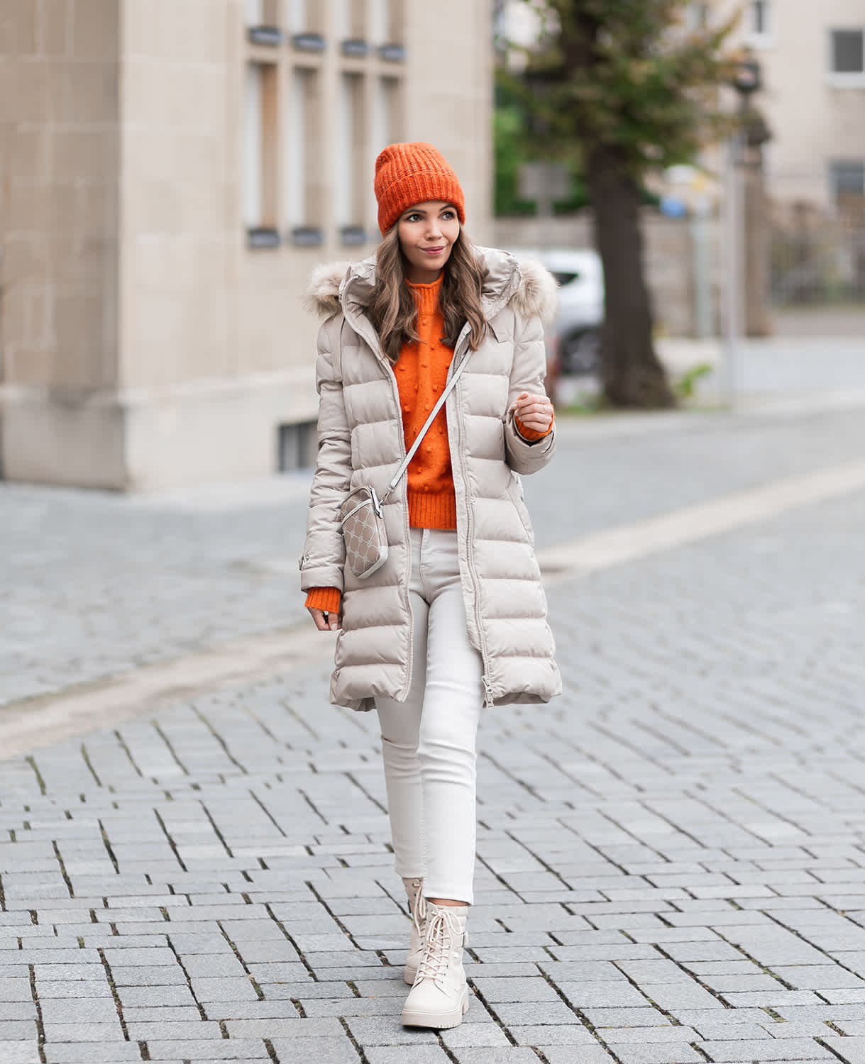 Frau in Winter-Outfit mit beigefarbenem Daunenmantel, orangem Pullover und Mütze, kombiniert mit hellen Boots