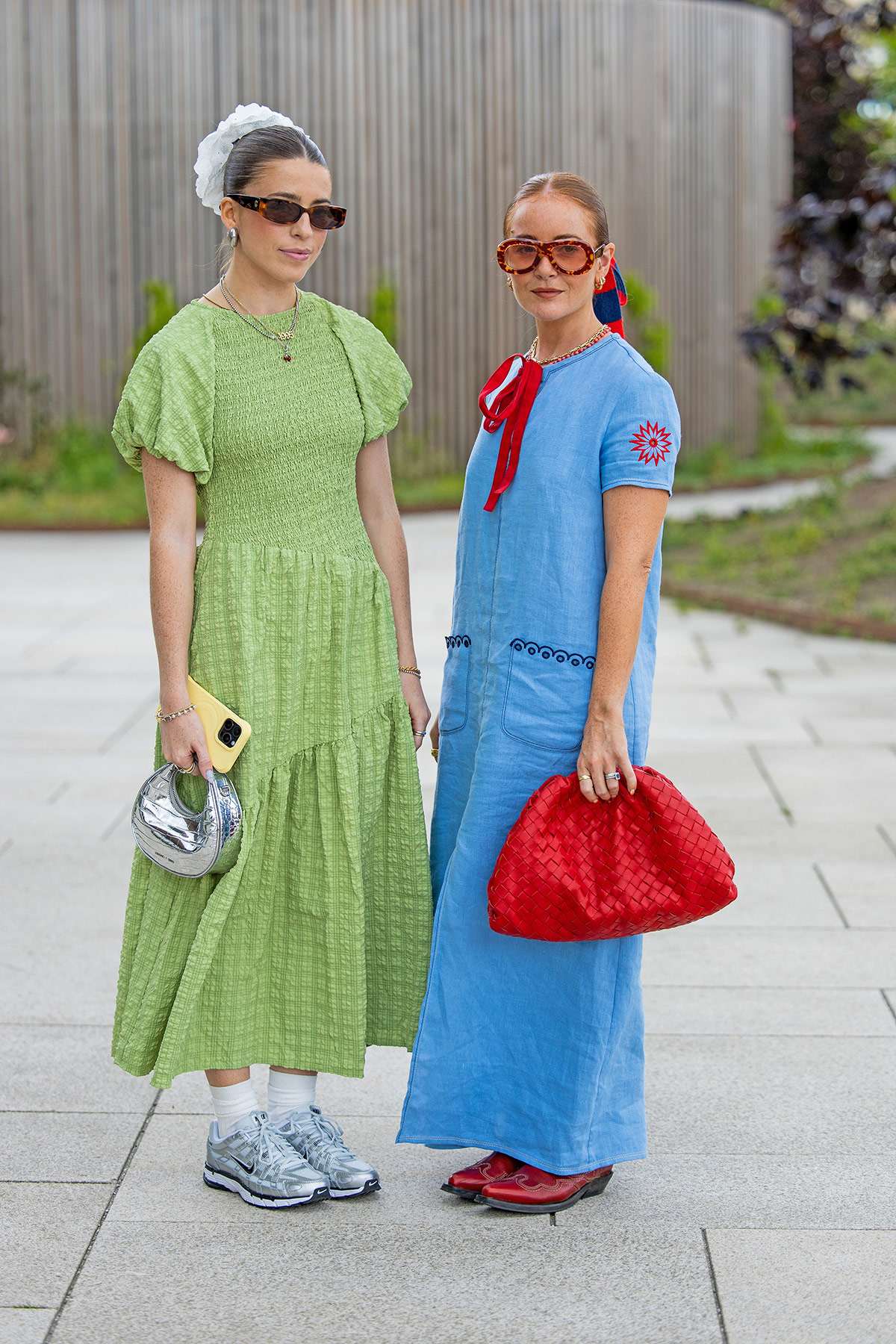Zwei Frauen mit Sonnenbrillen stehend nebeneinander, links in grünem Kleid, rechts in blauem Kleid mit roter Handtasche