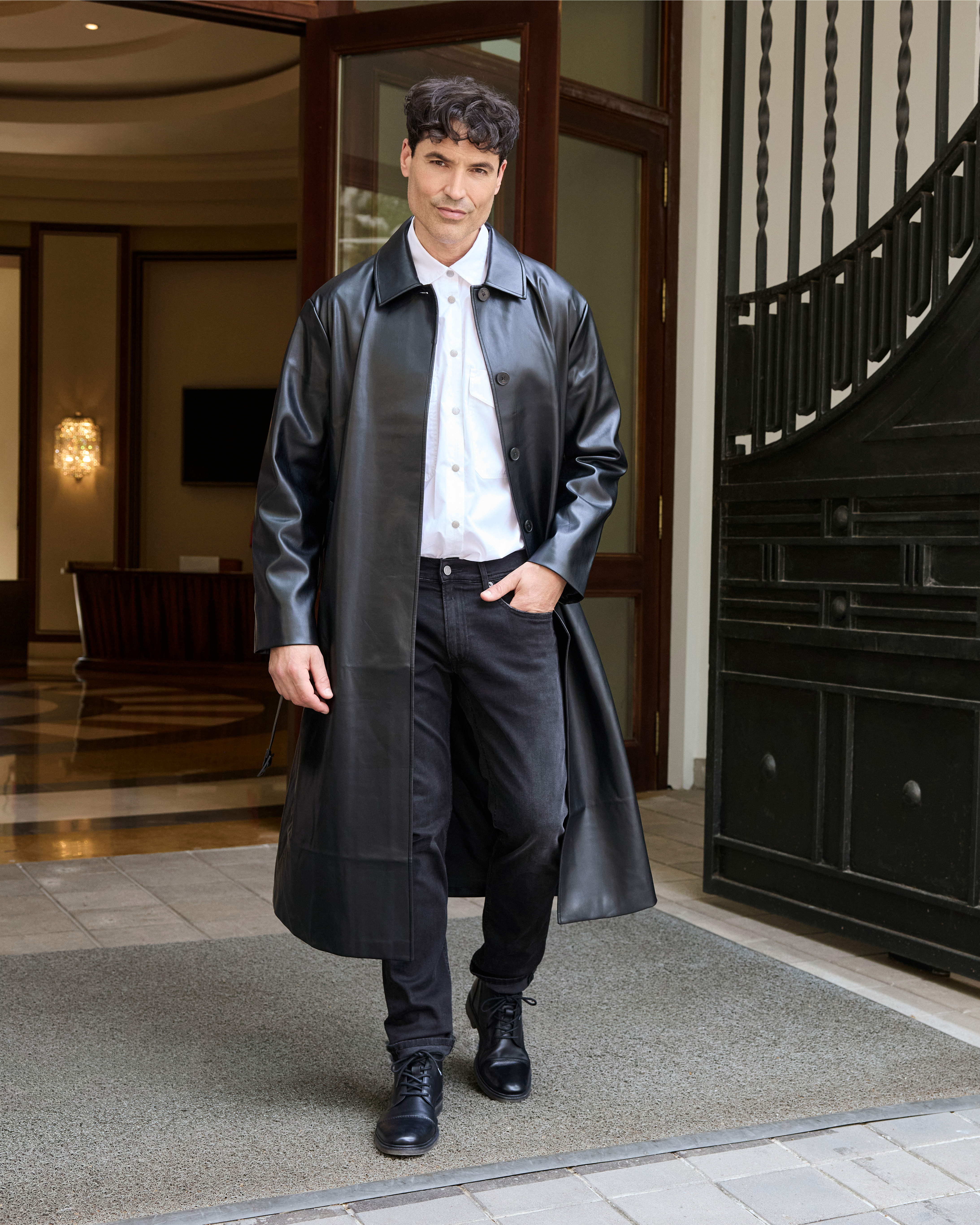 Un uomo indossa un lungo cappotto di pelle, una camicia bianca e dei jeans neri