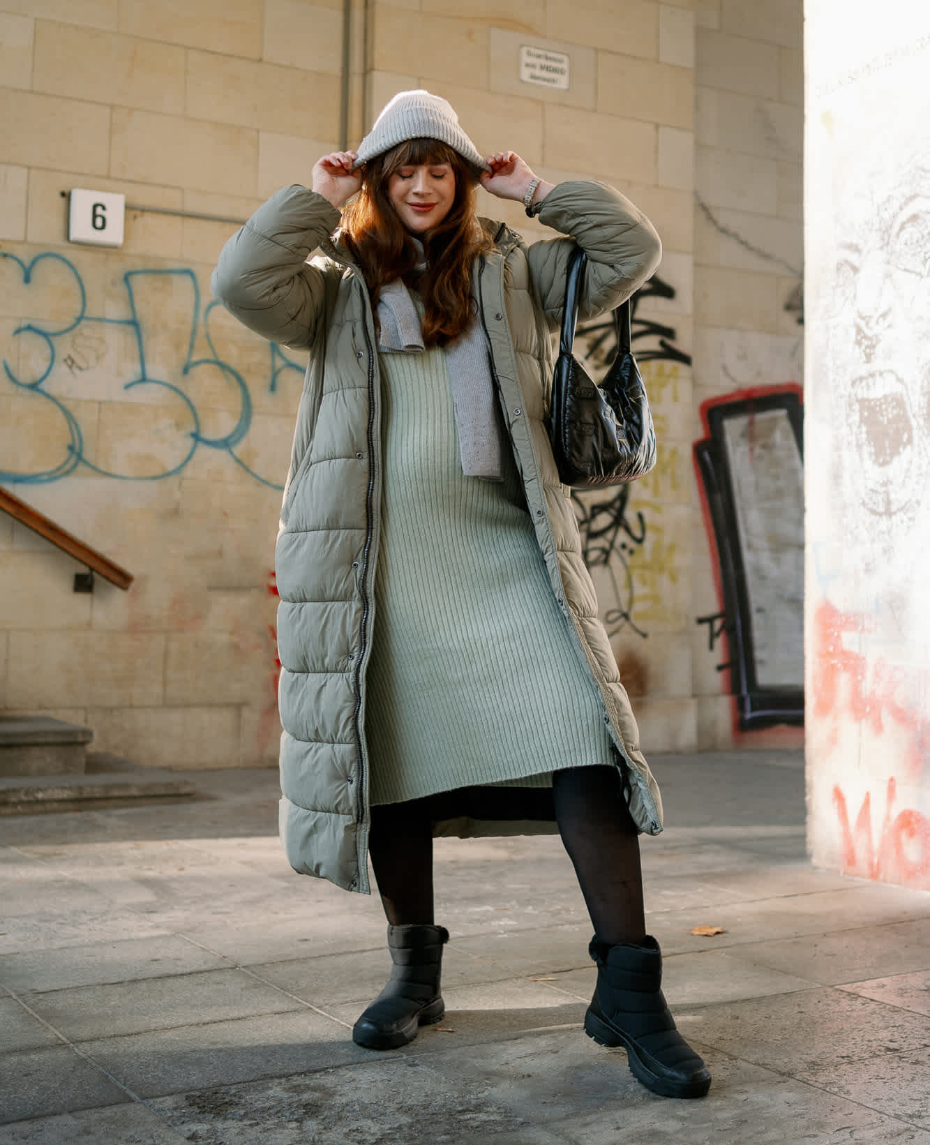 Frau mit offenem Oversize-Mantel in Hellgrün, mit farblich passendem Strickkleid, justiert mit beiden Händen Strickmütze auf Kopf