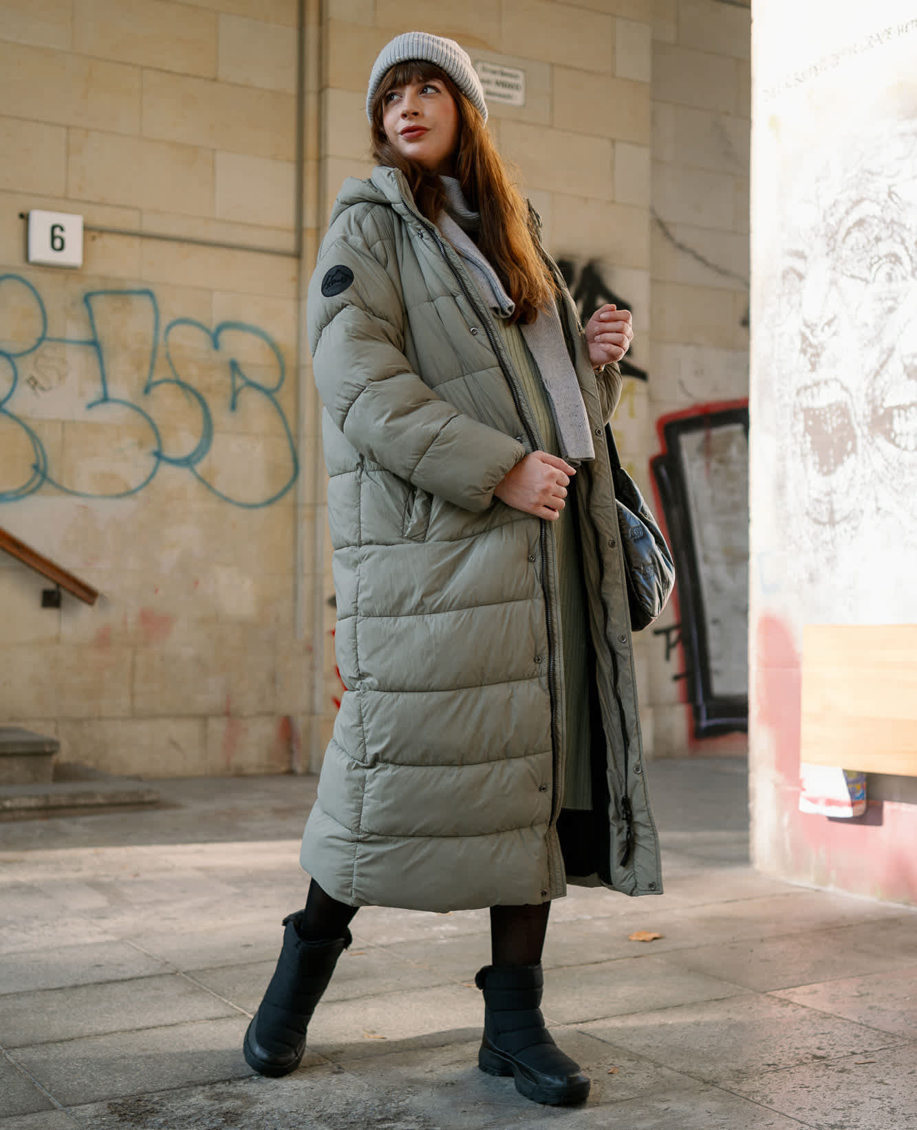 Frau in gestepptem Oversize-Mantel in Hellgrün, mit schwarzen Skechers-Stiefeln, entspannt stehend, mit Graffiti-Wand im Hintergrund