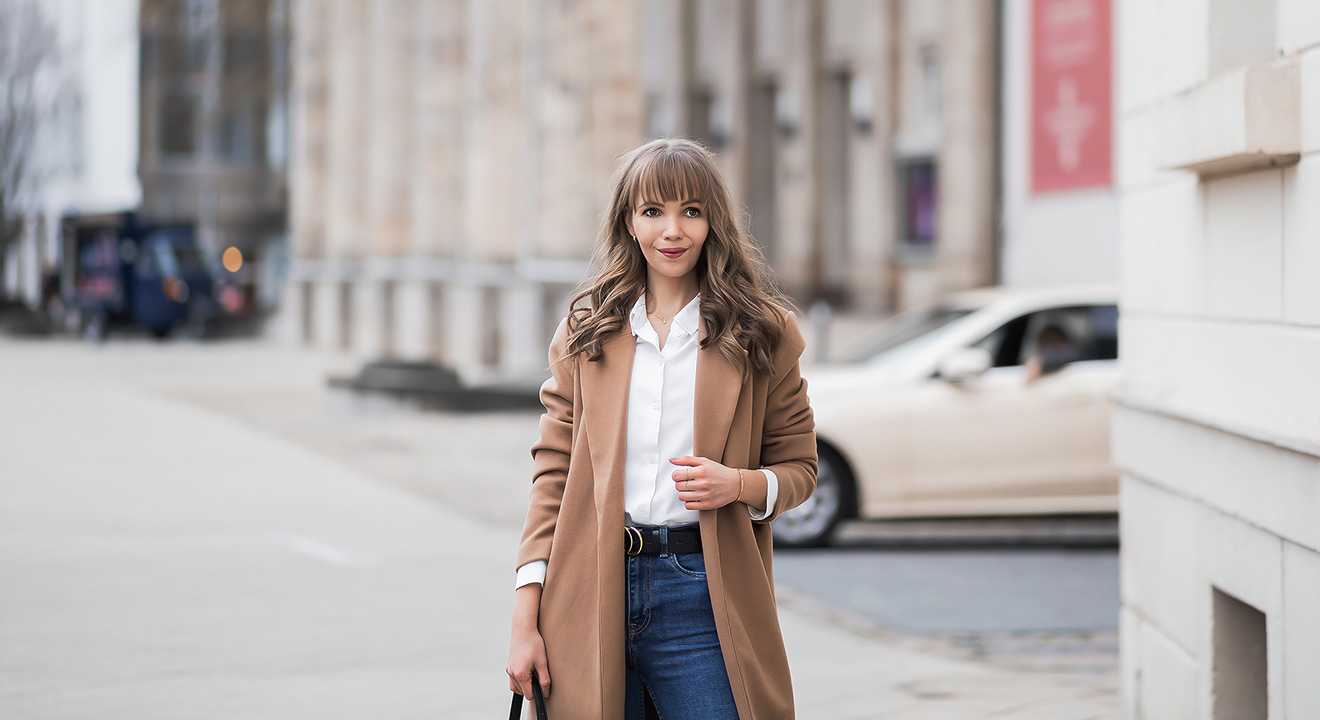 Frau in camel-farbenem Mantel, mit weißer Bluse und blauer Jeans, mit Handtasche in der Hand, vor urbaner Kulisse 
