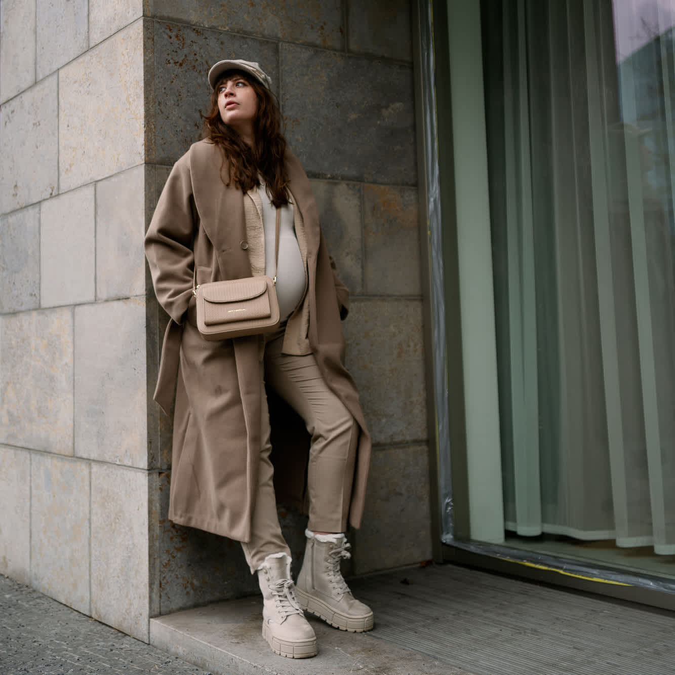 Frau in beigem Outfit samt Mantel und Handtasche lehnt entspannt an Gebäude