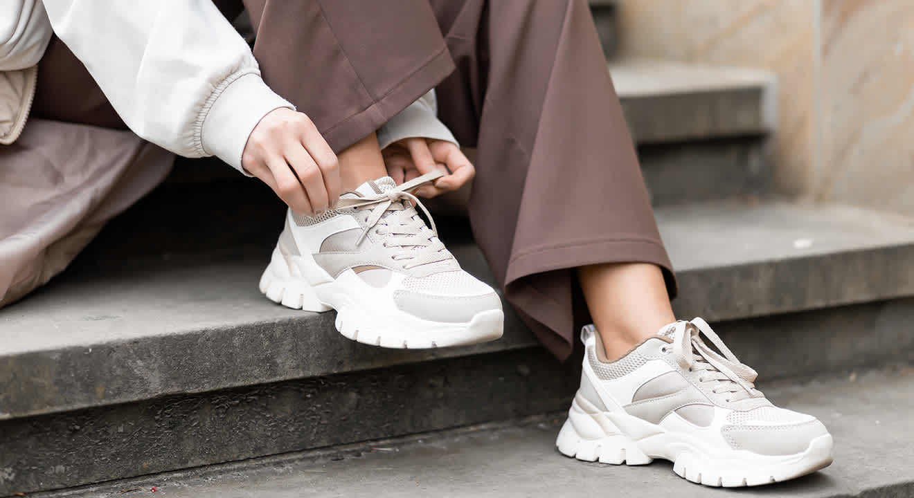 Person sitzt auf Steinstufen und bindet beigefarbene Sneaker, getragen zu einer braunen, weiten Hose und cremefarbenem Sweatshirt.
