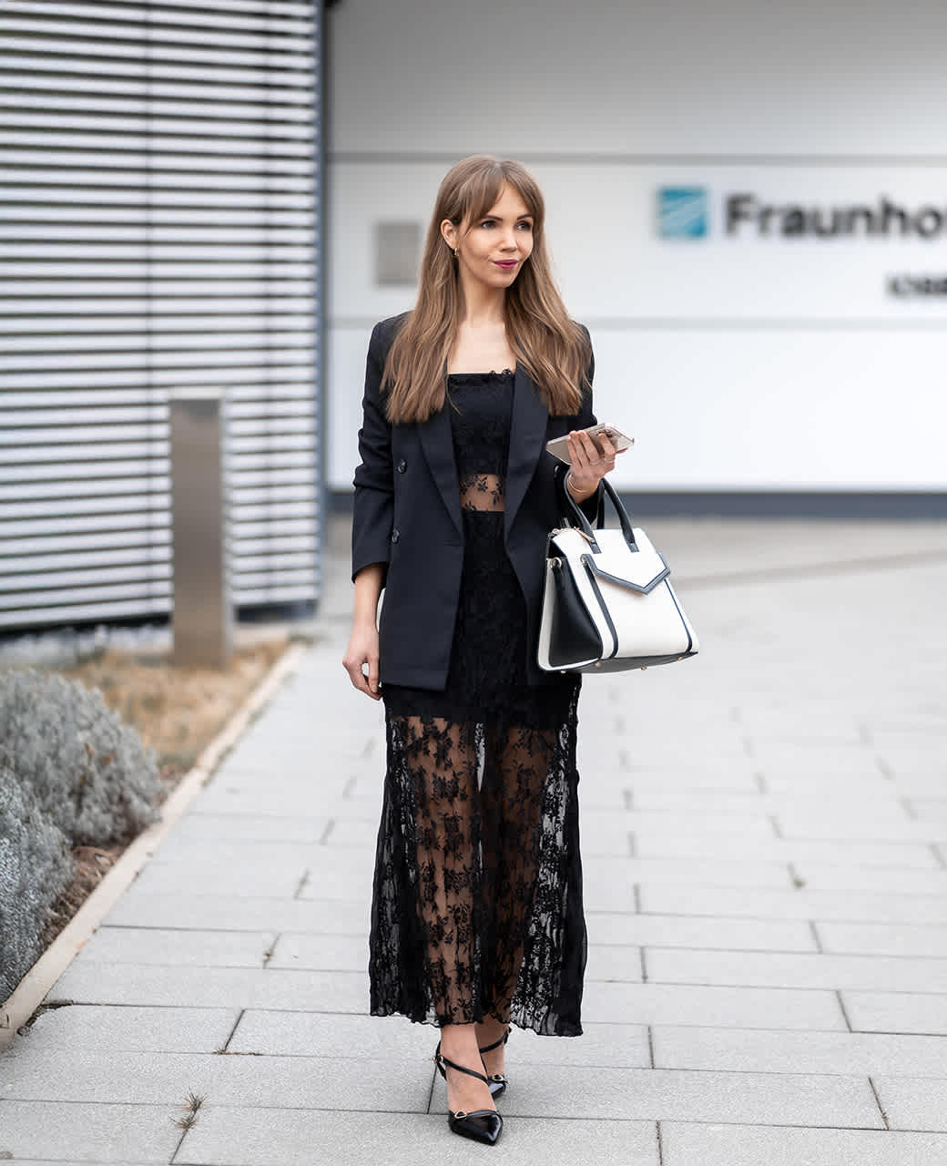 Frau geht über einen Gehweg vor einem modernen Gebäude, trägt ein schwarzes Spitzenkleid mit Blazer und hohe Schuhe. In der Hand hält sie eine große schwarz-weiße Handtasche.