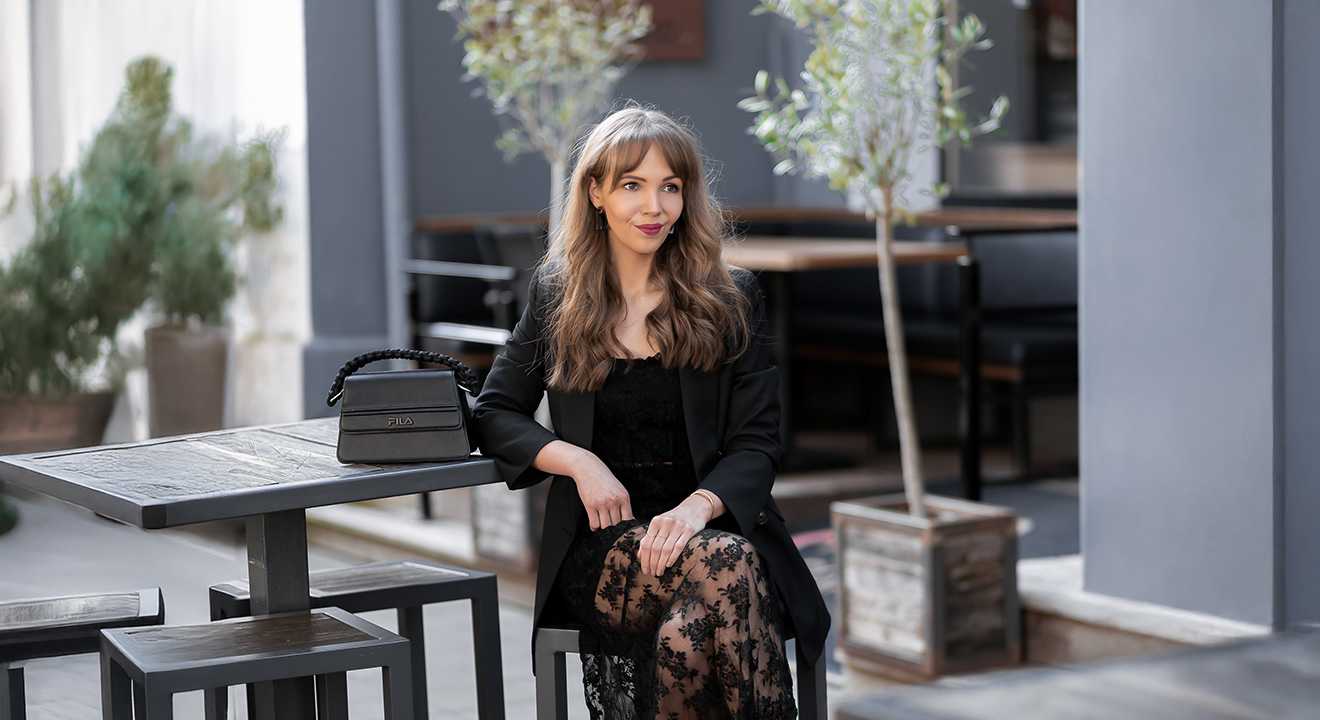 Frau sitzt an einem Tisch im Außenbereich eines Cafés, trägt ein schwarzes Spitzenkleid mit Blazer und hat eine schwarze Handtasche von Fila auf dem Tisch.