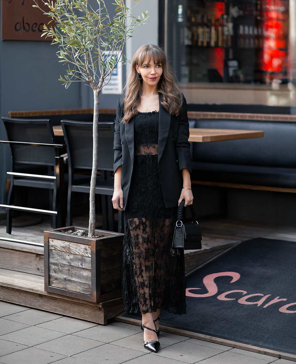 Frau steht vor einem Restaurant in schwarzem Spitzenkleid mit Blazer und Absatzschuhen. In der Hand trägt sie eine kleine schwarze Handtasche.