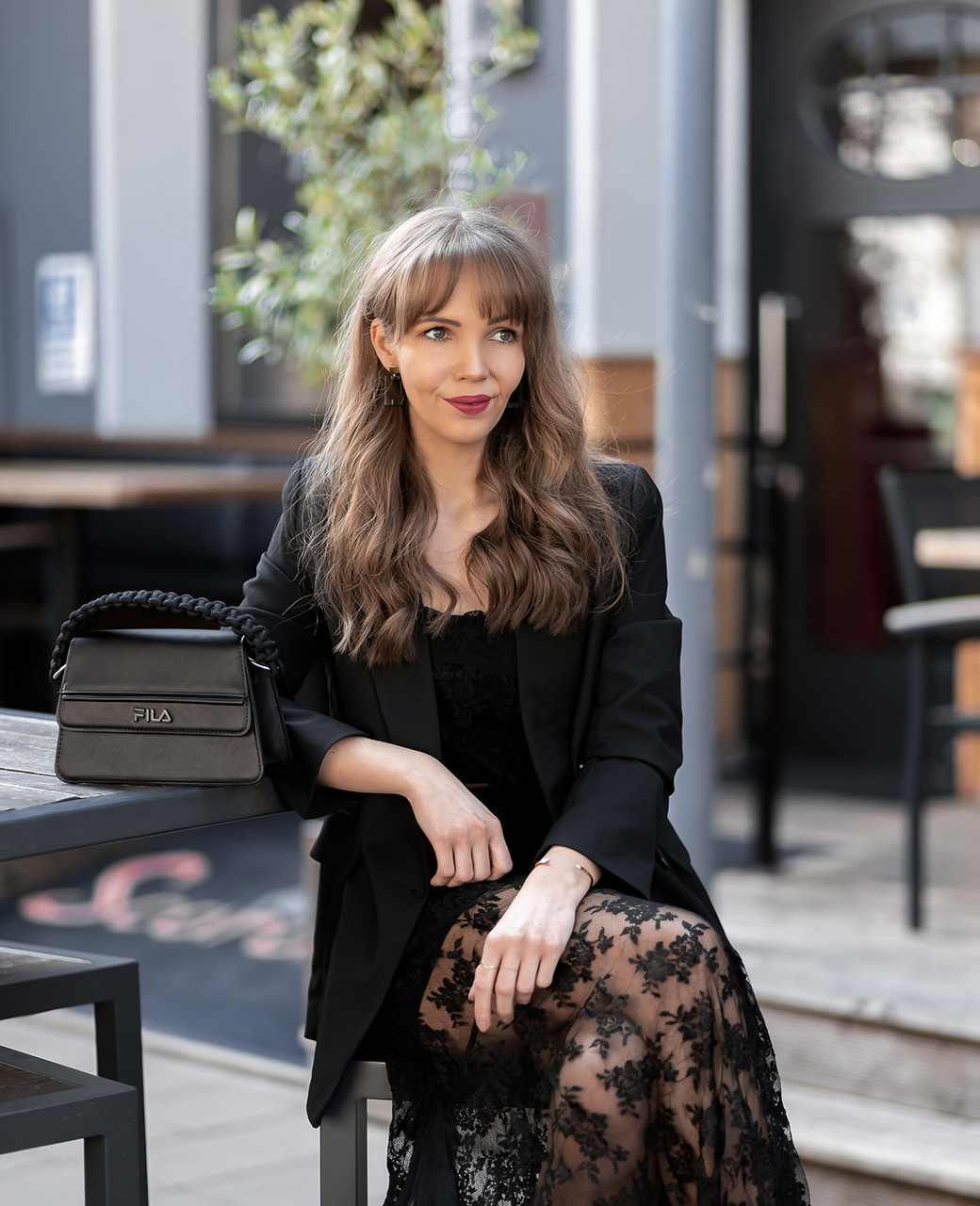 Frau sitzt im Außenbereich eines Cafés in schwarzem Spitzenkleid mit Blazer. Auf dem Tisch neben ihr steht eine schwarze Handtasche von Fila.