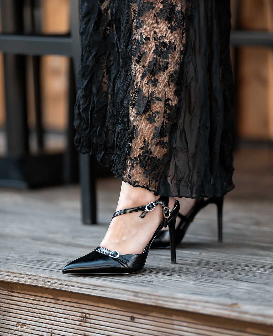 Nahaufnahme von schwarzen Riemchen-High Heels mit spitzer Kappe unter einem langen, schwarzen Spitzenkleid.