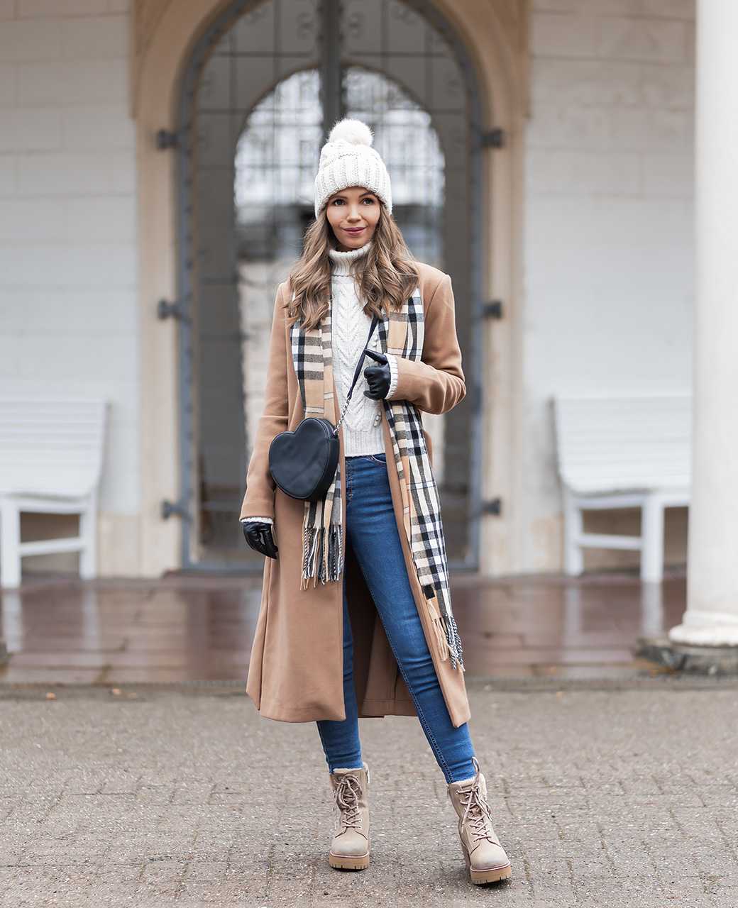 Frau steht in Winter Outfit vor Gebäude, trägt braunen Mantel, modisch-karierten Schal, blaue Jeans und Tom Tailor-Stiefeletten