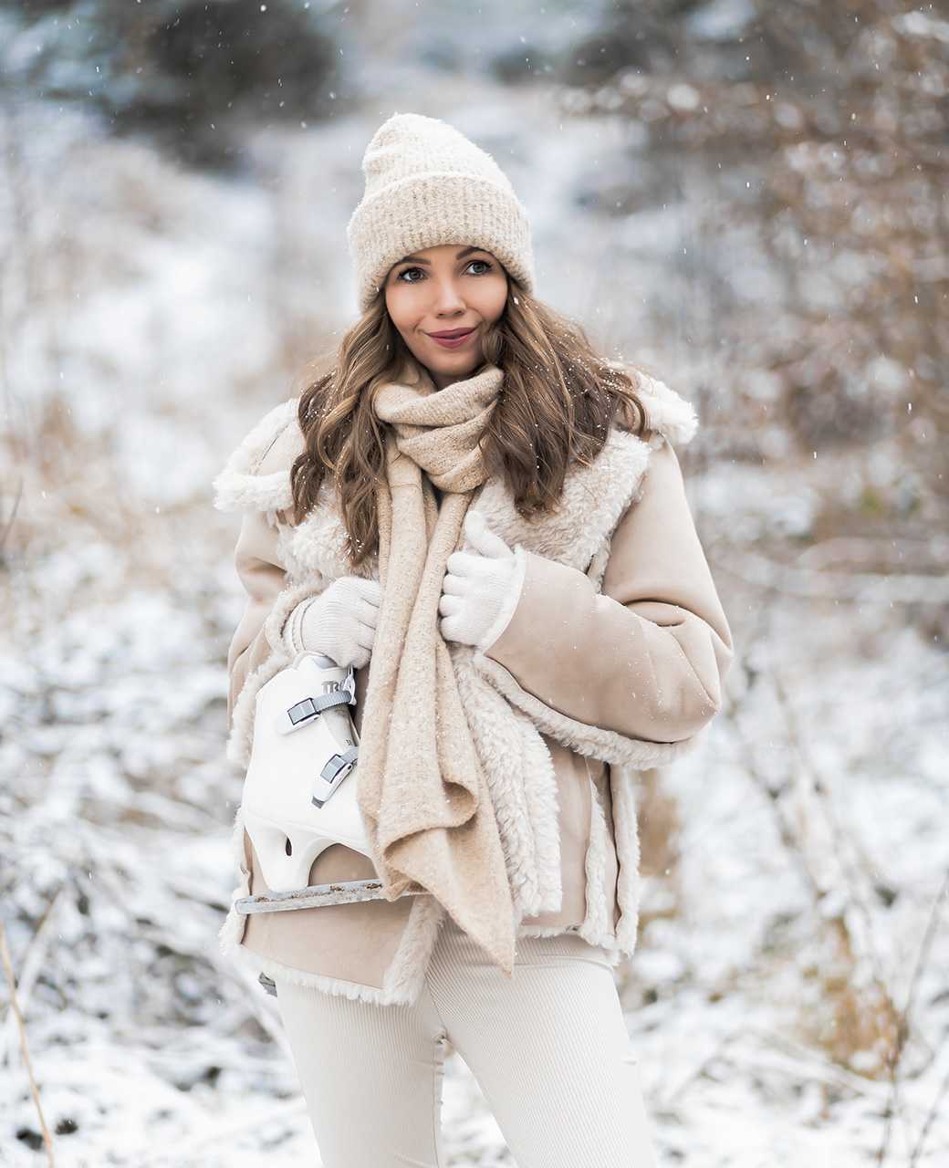 Frau trägt kuscheliges Winter Outfit aus beiger Jacke mit Kunstfell, Strickschal & -mütze, zusammen mit cremefarbener Hose