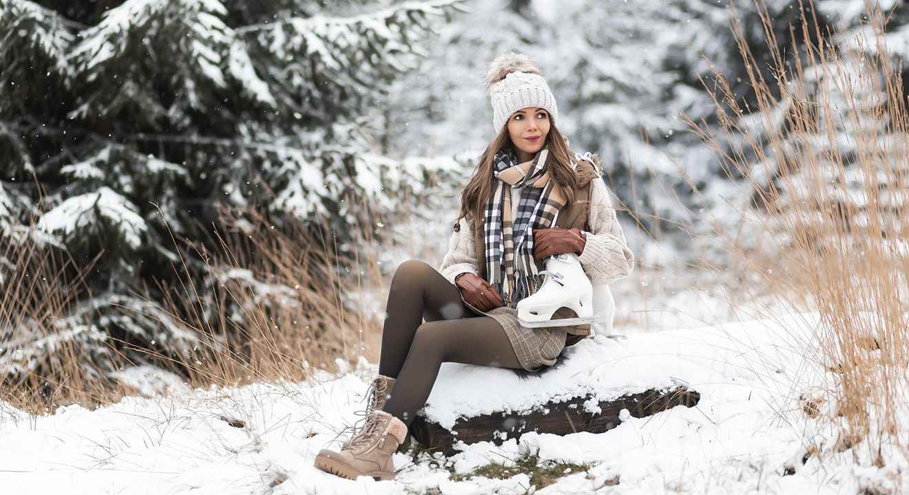 Frau sitzt auf Baumstamm in Winterlandschaft, trägt warme Kleidung und ein Paar weiße Schlittschuhe am Arm