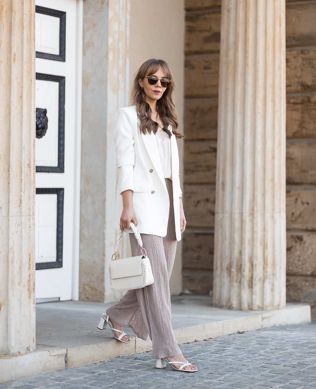 Frau in elegant weißem Outfit, spaziert lässig vor Steinfassade, mit weißer Handtasche