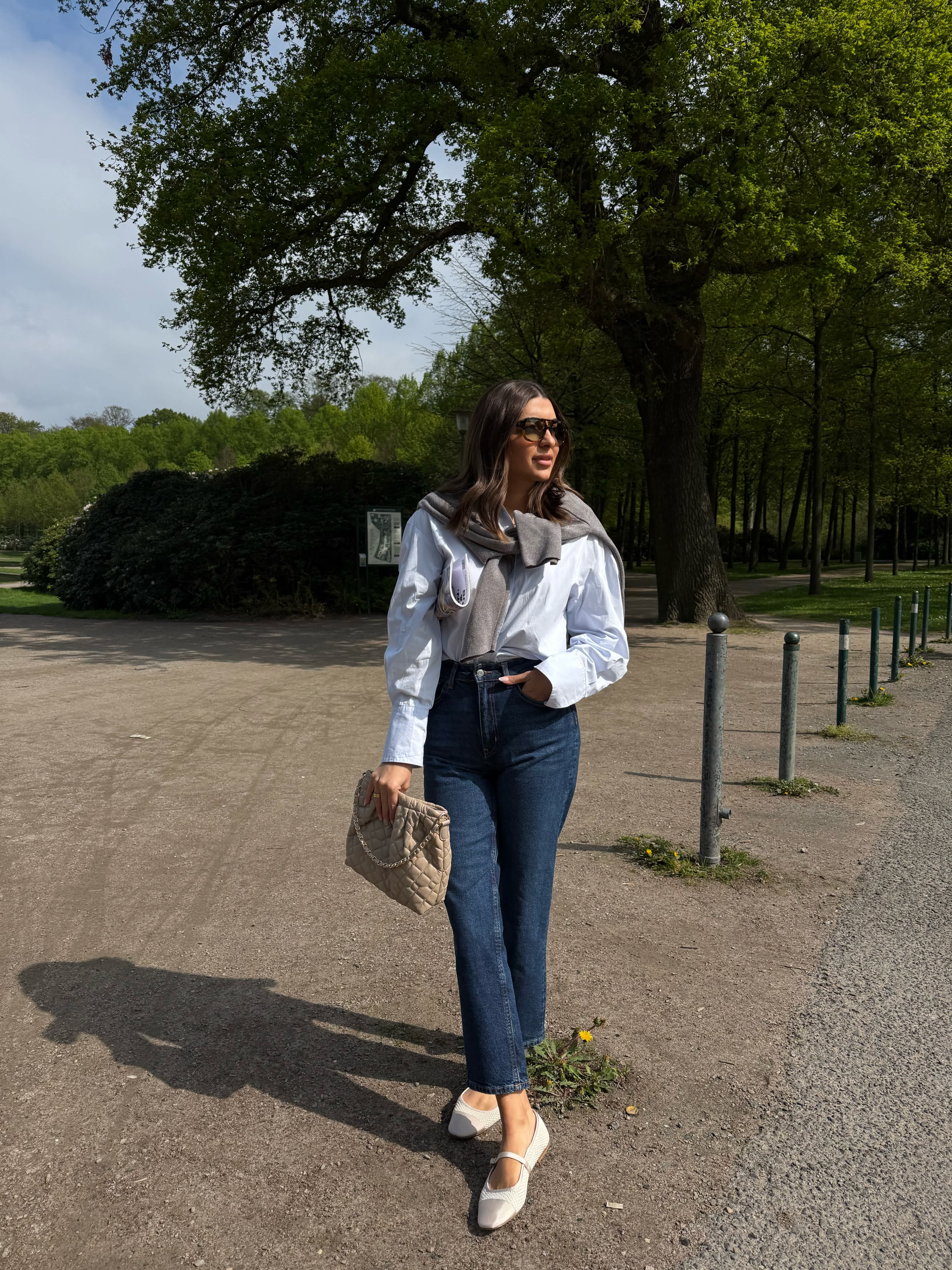 Frau steht in lässigem Business-Outfit im Park, trägt Kombination aus Jeans und weißer Bluse