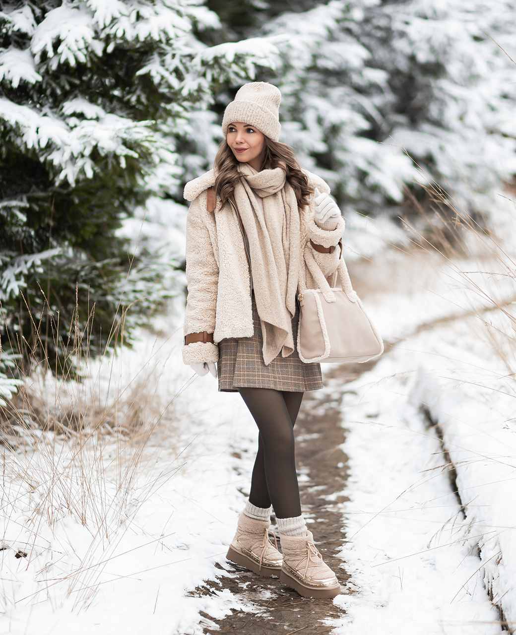 Frau ist gekleidet in beigefarbenes Outfit aus Fellimitat, trägt passende Handtasche und steht auf verschneitem Feldweg