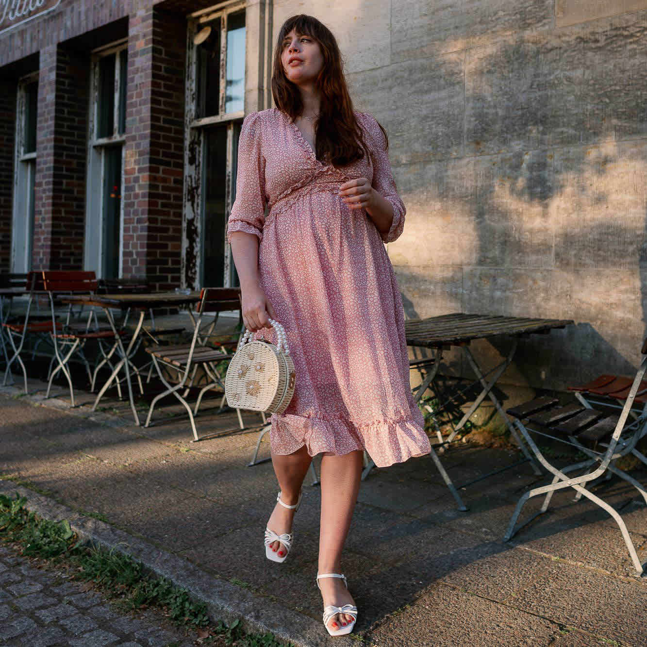 Frau in pastellfarbenem Kleid, kombiniert weiße Sandalen mit Bast-Tasche