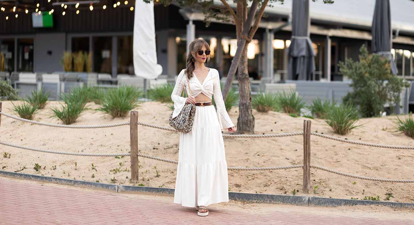 Frau in Boho-Outfit mit weißem Top und passender luftiger Hose, vor Strandcafé-Kulisse