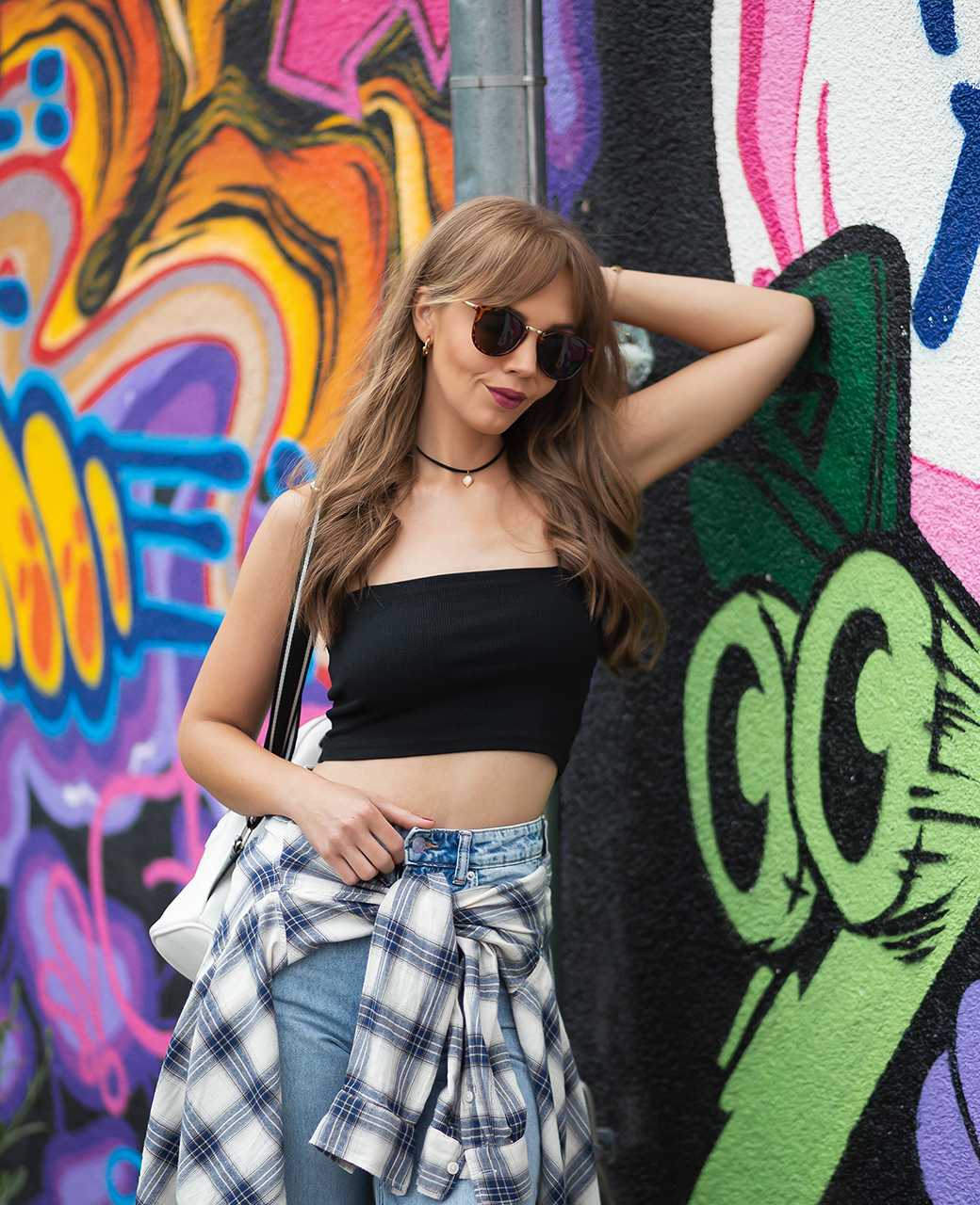 Frau trägt Y2K-Style-Outfit mit schwarzem Top und blauer Jeans, lehnt an bunter Wand 