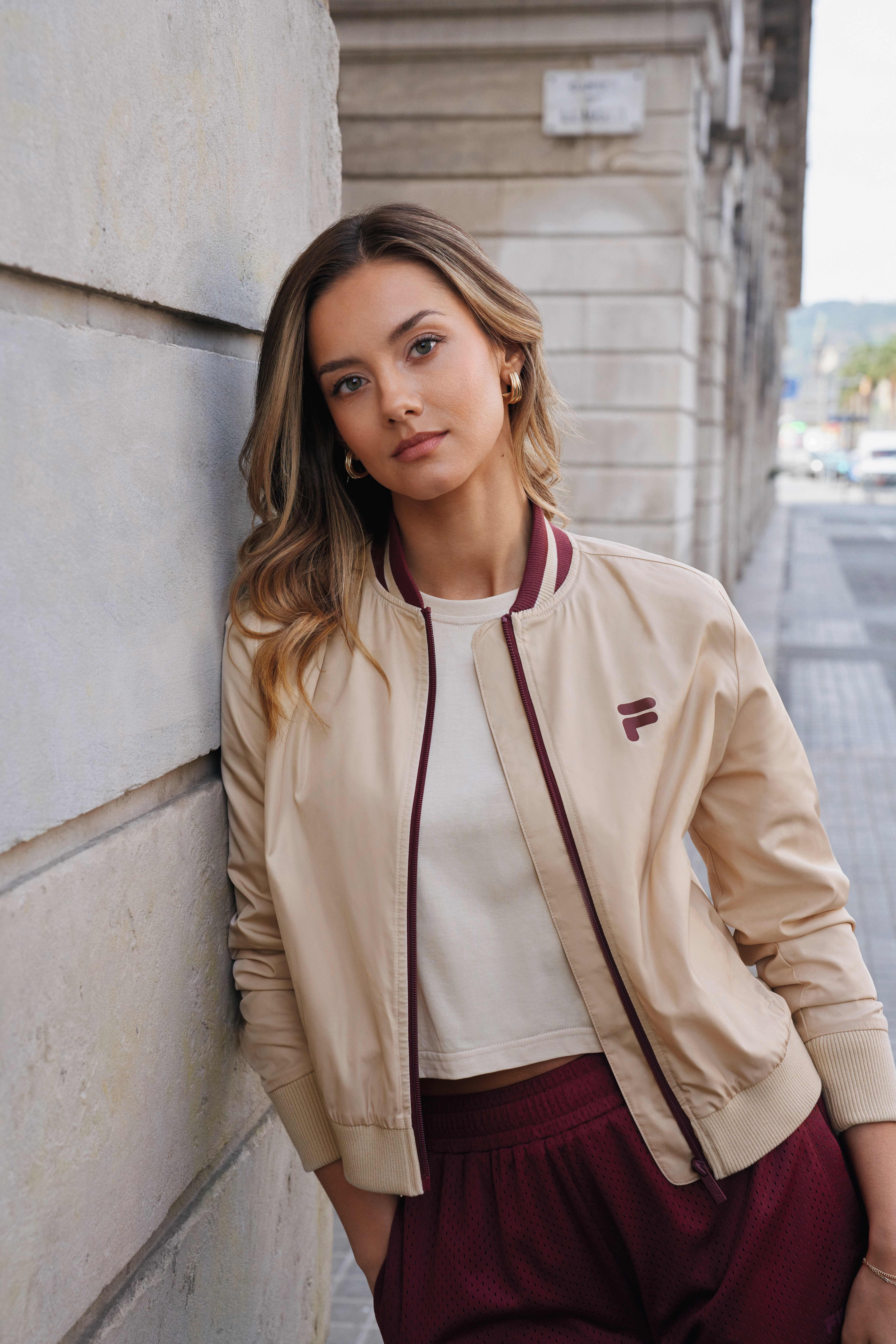 Eine Frau trägt ein weißes Crop-Top mit einer beigefarbenen Bomberjacke und weinroten Shorts