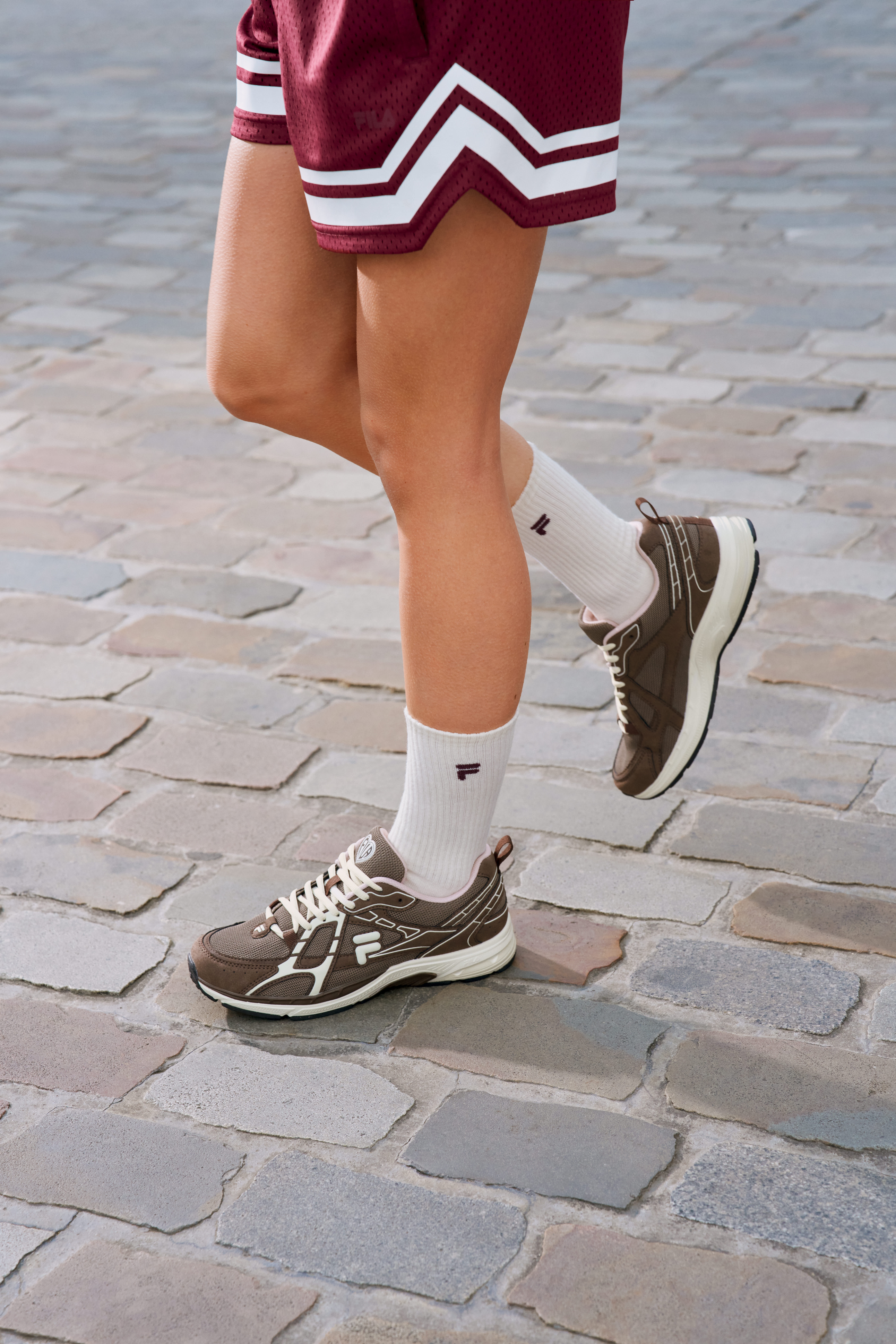 Eine Frau trägt ein weinrotes Sportset mit weißen Streifen, dunkelbraune FILA-Sneaker und weiße Socken