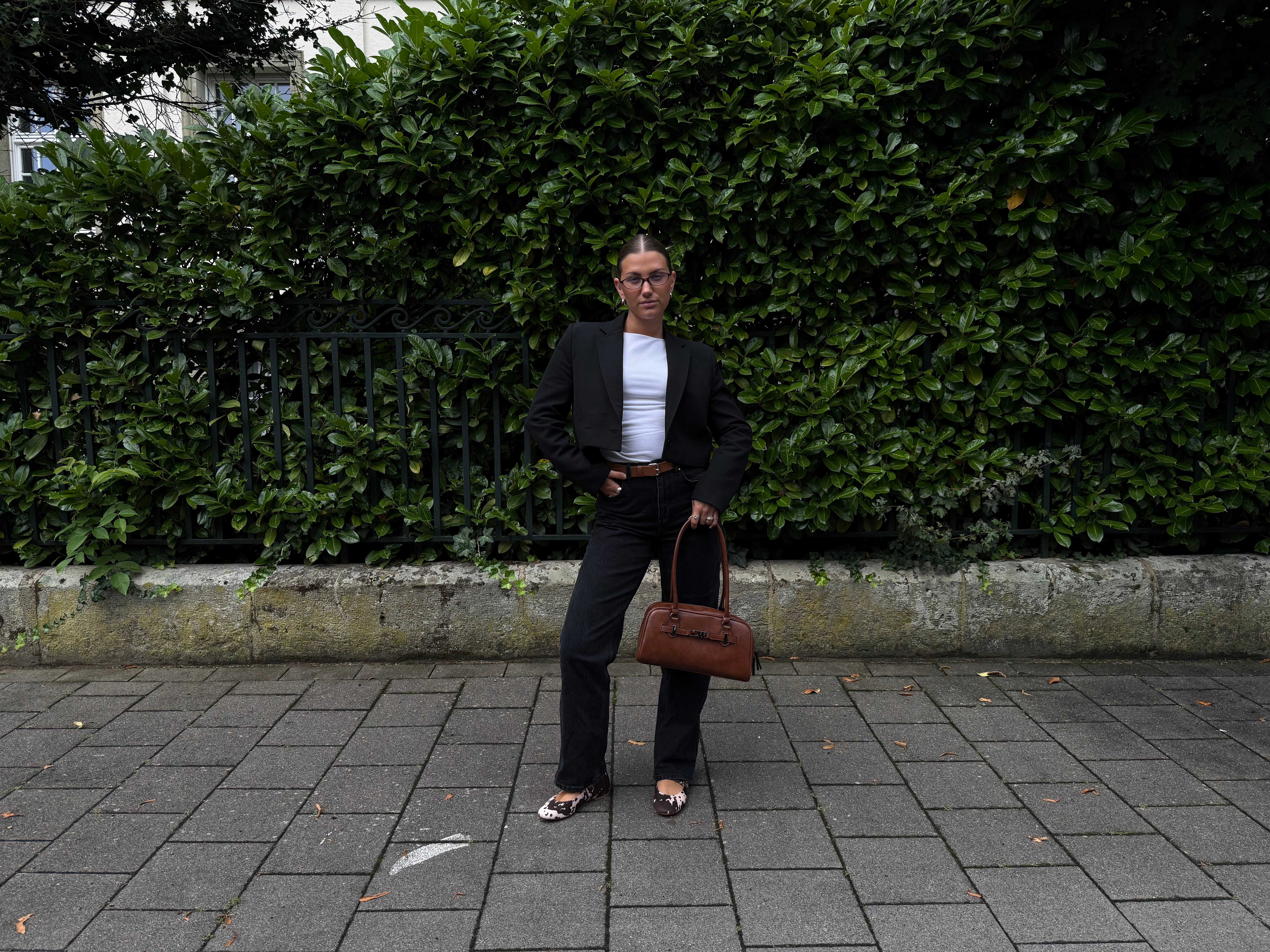 Frau mit dunklem Blazer und passender Hose, kombiniert mit weißem Shirt und brauner East West Bag