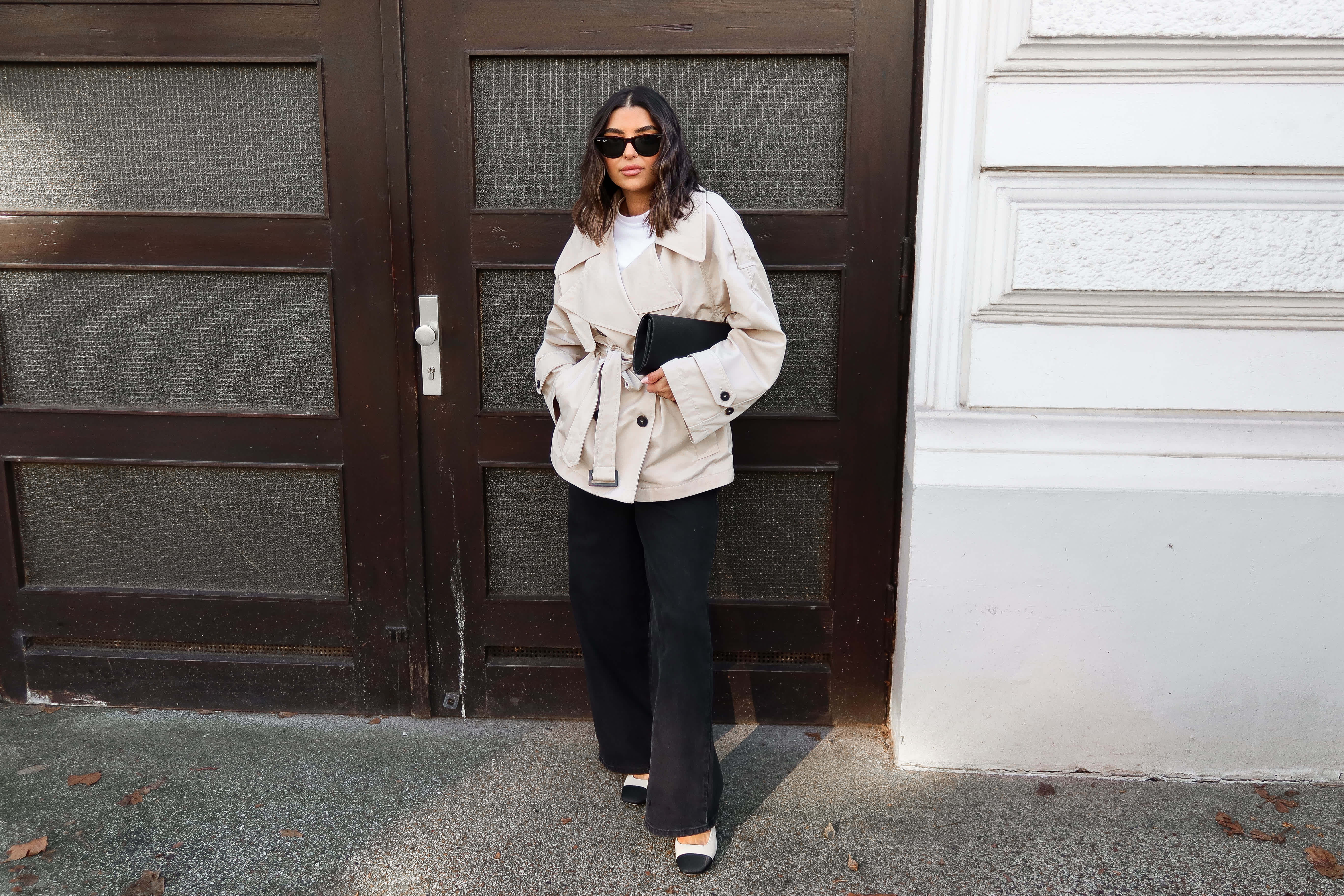 Frau mit beigem Trenchcoat, schwarzer weiter Hose und passender Clutch steht in einem Hauseingang