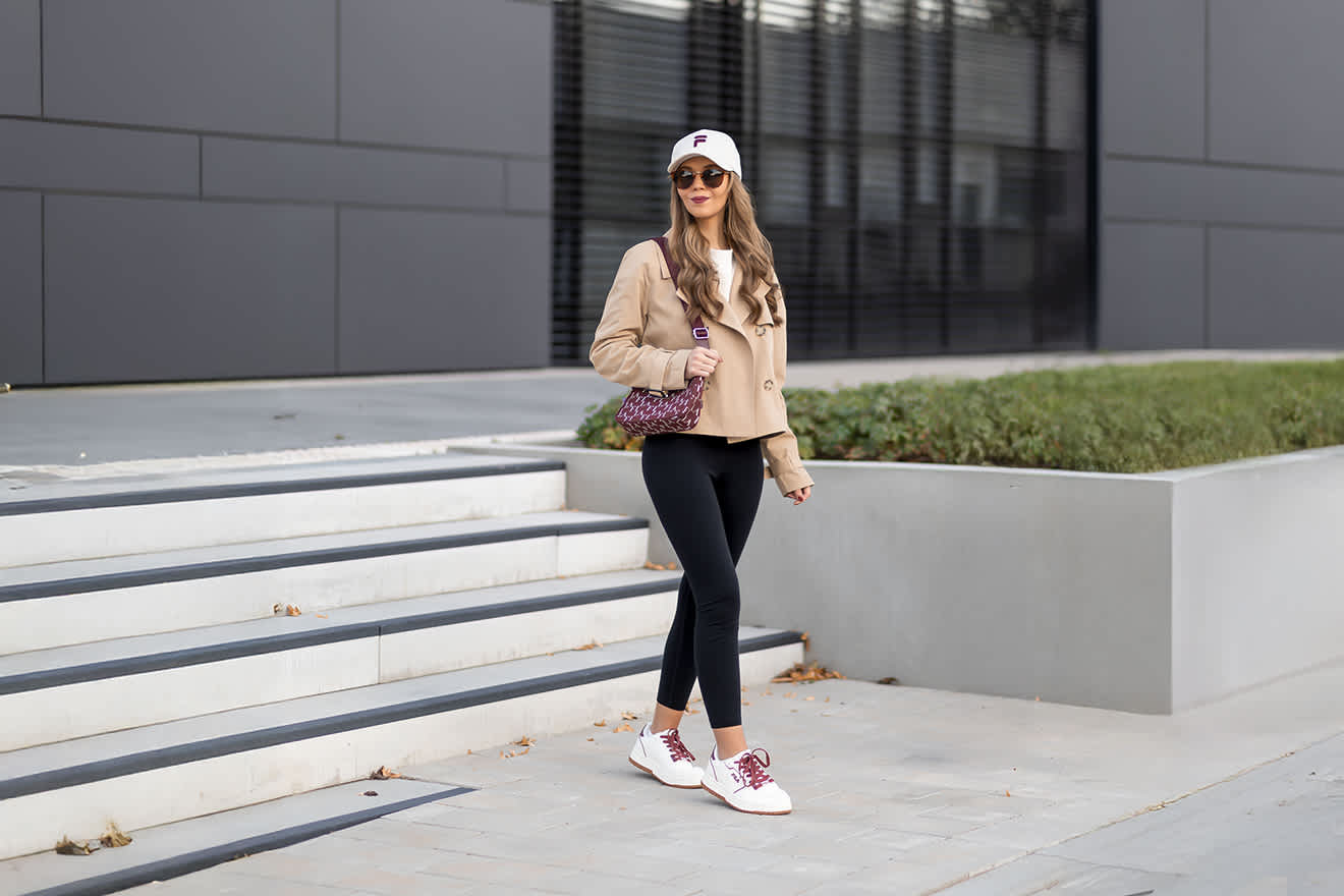 Frau in braunem Cropped Trenchcoat und schwarzer Leggings, mit Handtasche und FILA-Cap