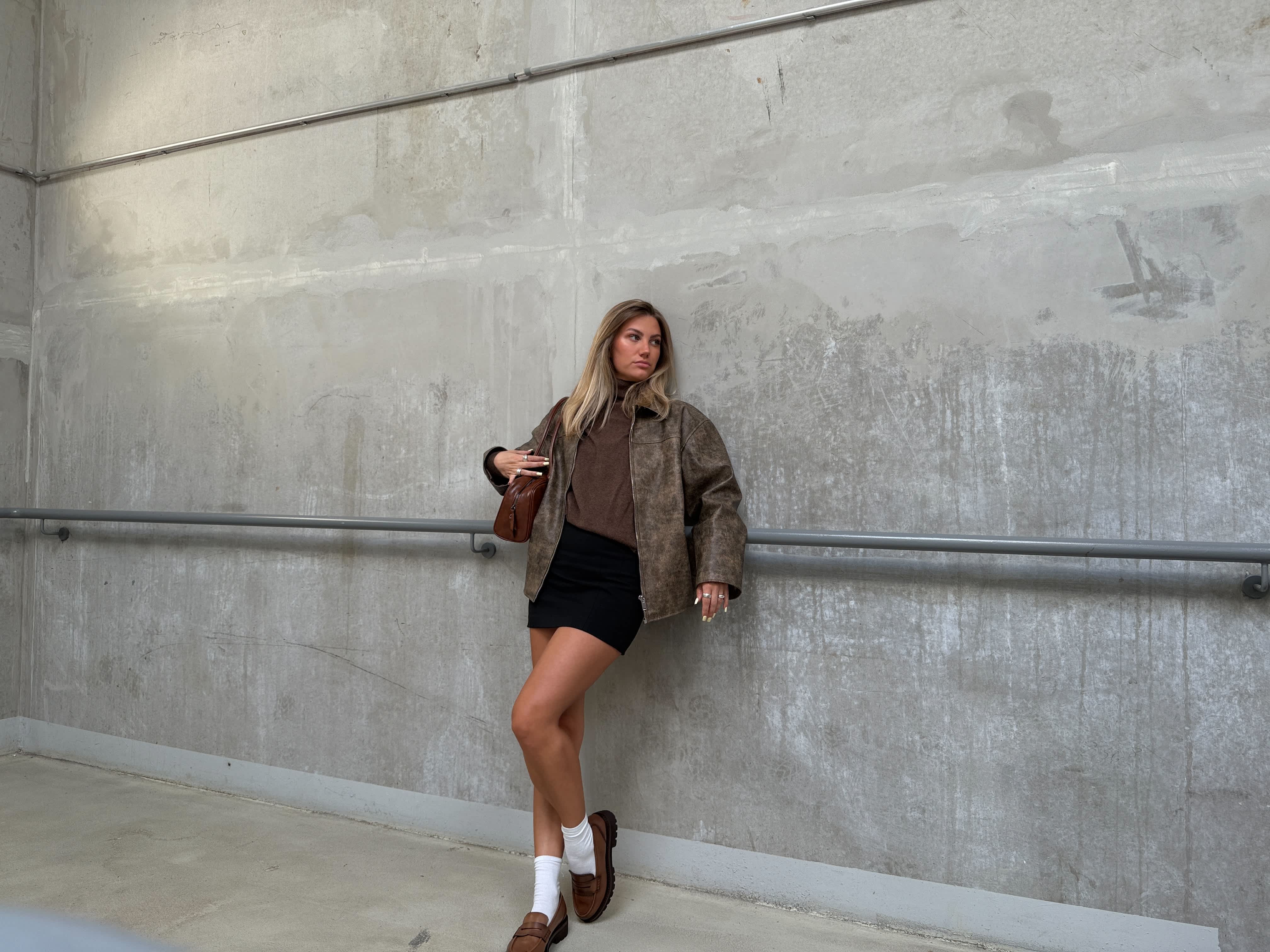 Frau in brauner Jacke, mit passendem Top und schwarzen Shorts, lehnt an Hauswand