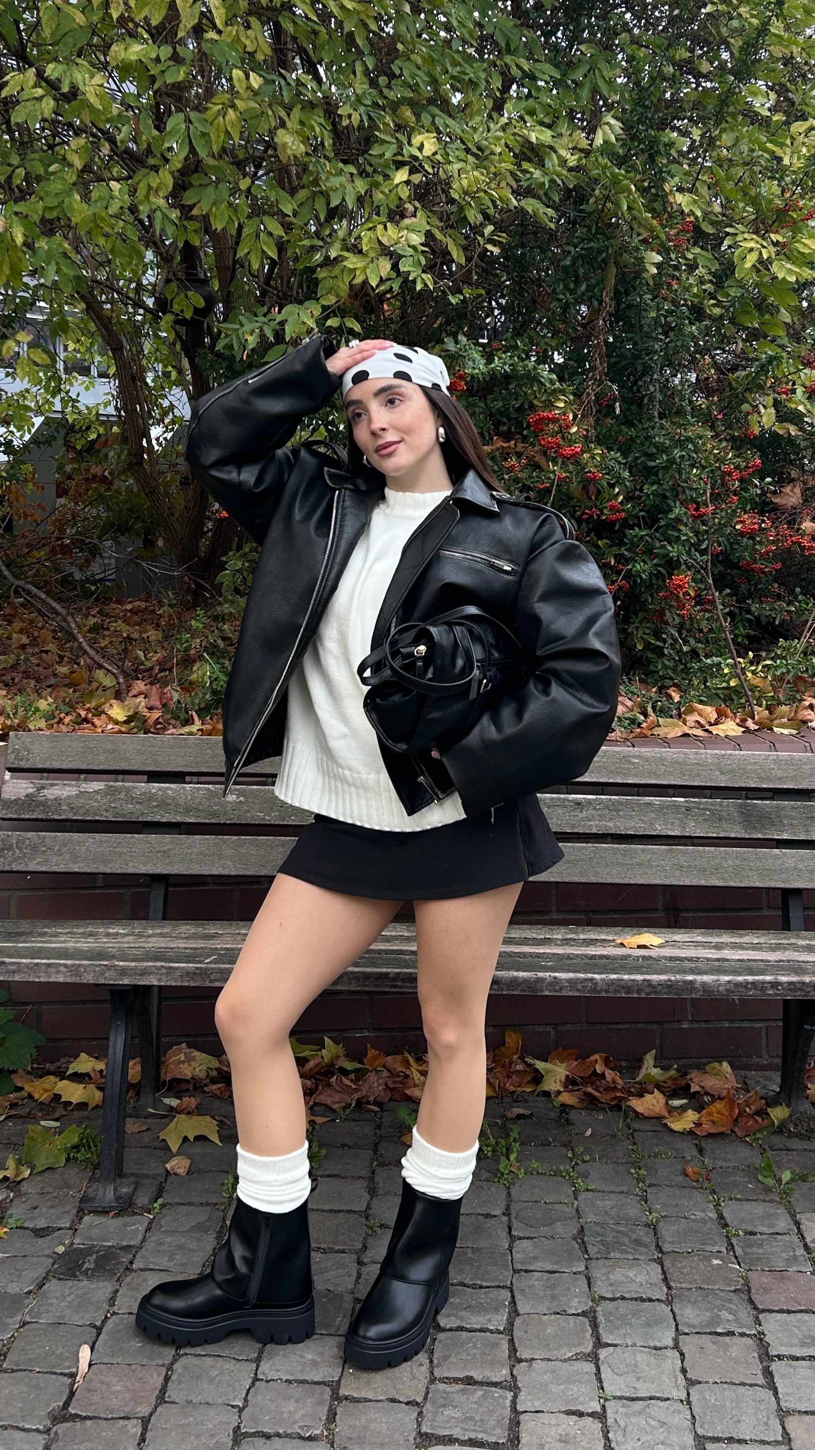 Junge Frau posiert in Park, trägt schwarze Jacke mit weißem Top, dazu schwarze Boots und Rock