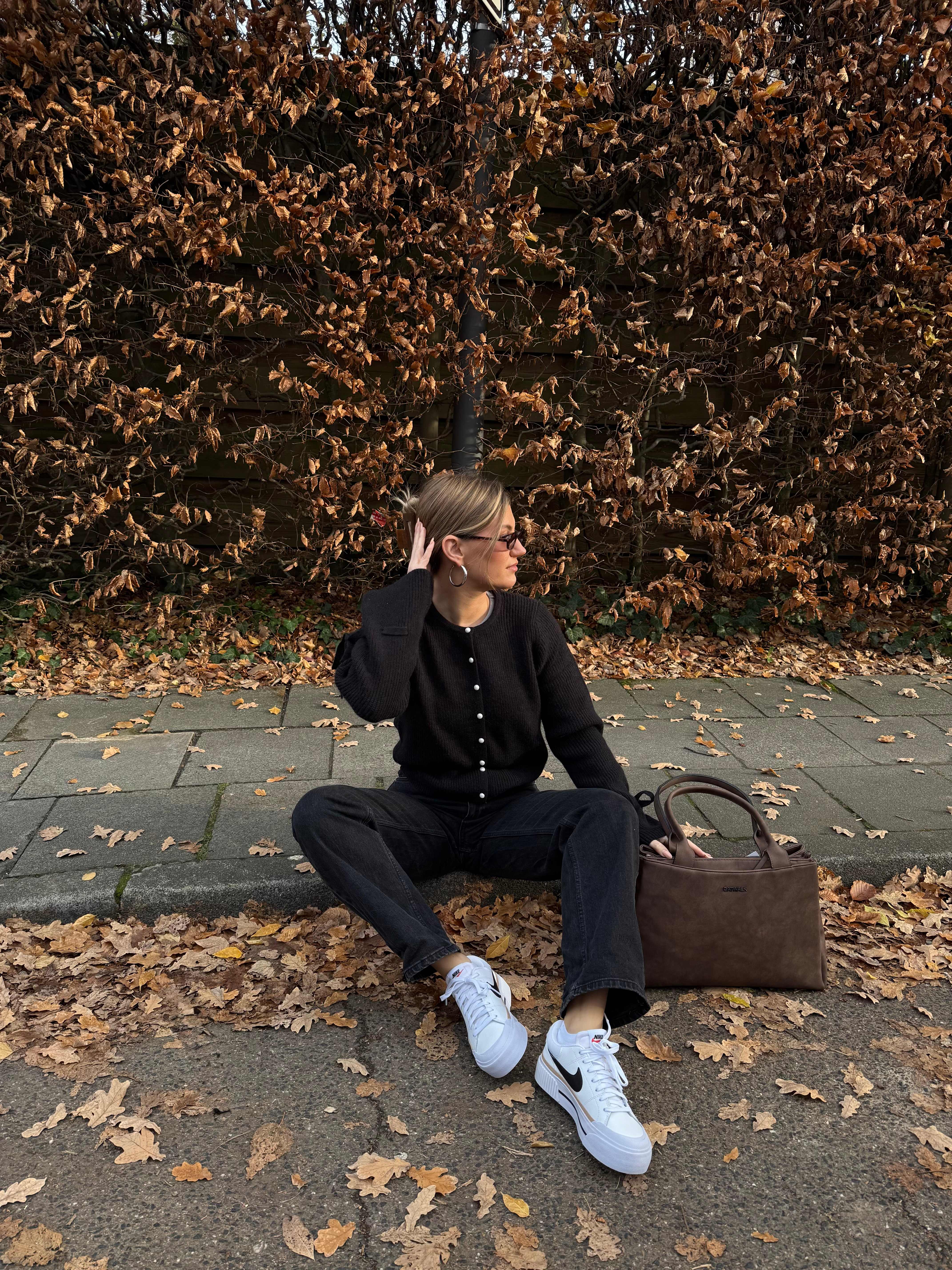 Frau in schwarzem Cardigan, kombiniert mit Hose und weißen Sneakern, sitzt am Straßenrand