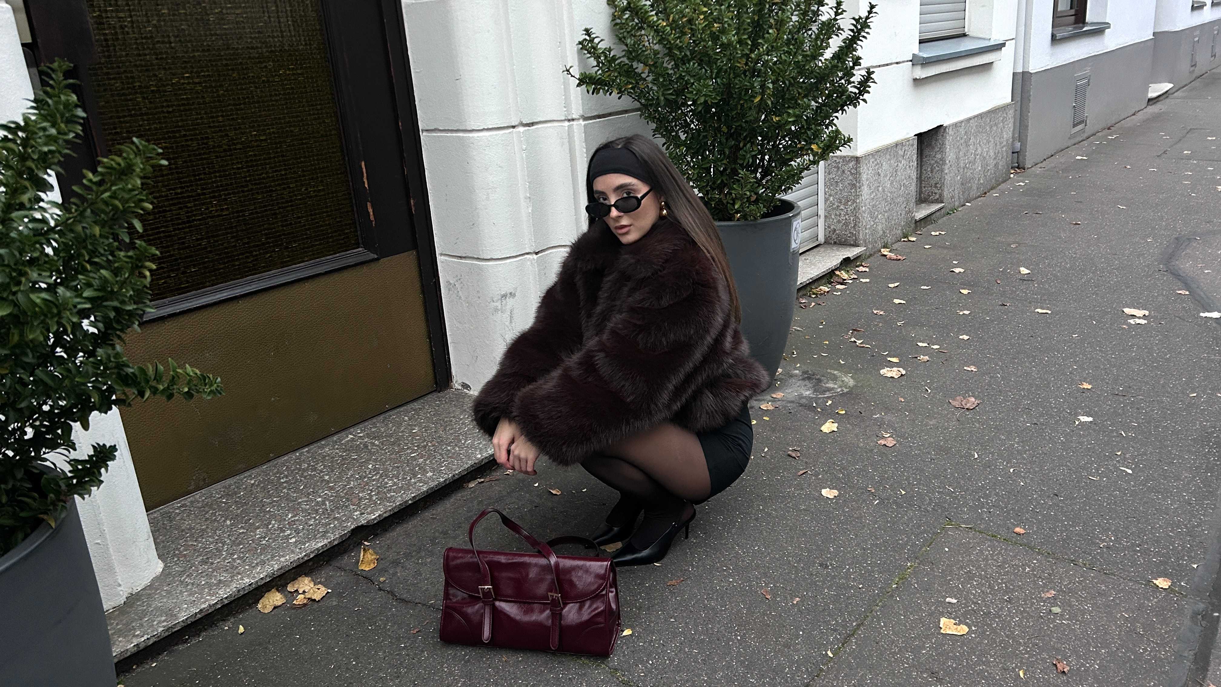 Frau im Mob Wife-Look, in kniender Position, mit roter Handtasche vor ohr