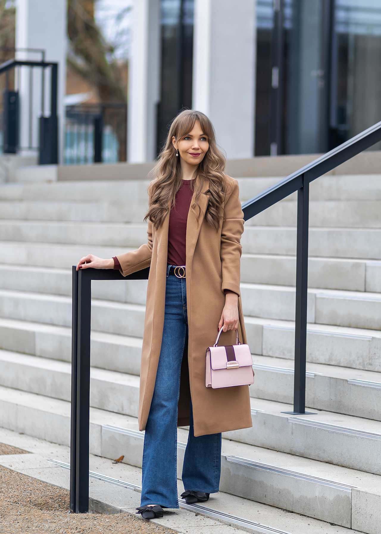 Junge Frau in braunem Mantel, mit Jeanshose und rosa Handtasche, an Treppengeländer