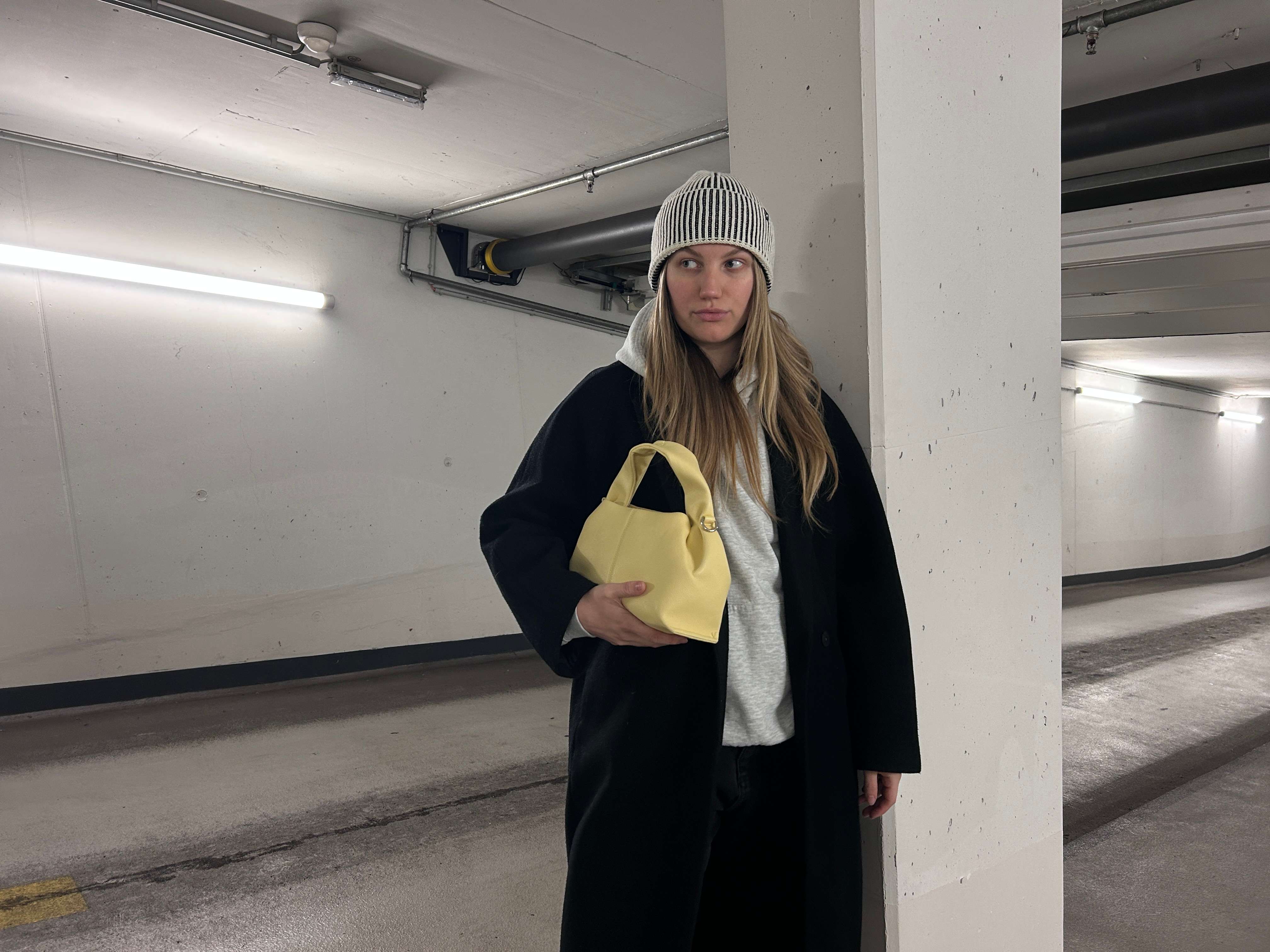 Frau trägt schwarzen Mantel und grauen Beanie, hält gelbe Handtasche unterm Arm