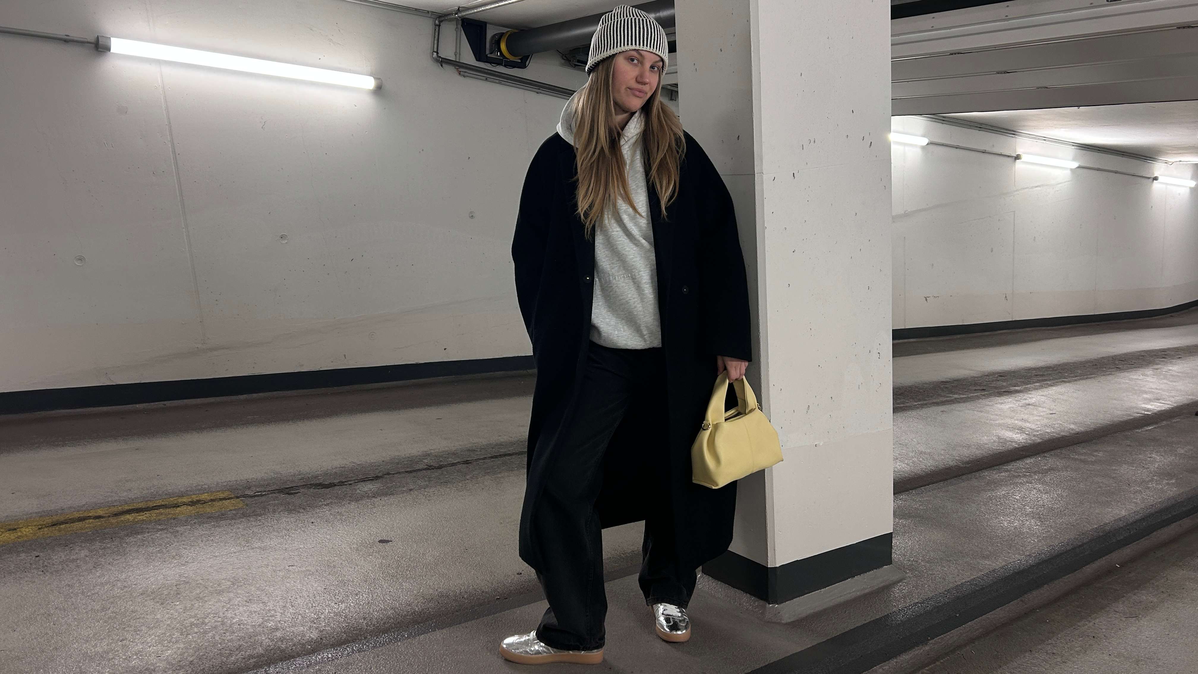 Frau in schwarzem Mantel und grauem Hoodie, trägt Metallic-Sneaker, steht in Tiefgarage