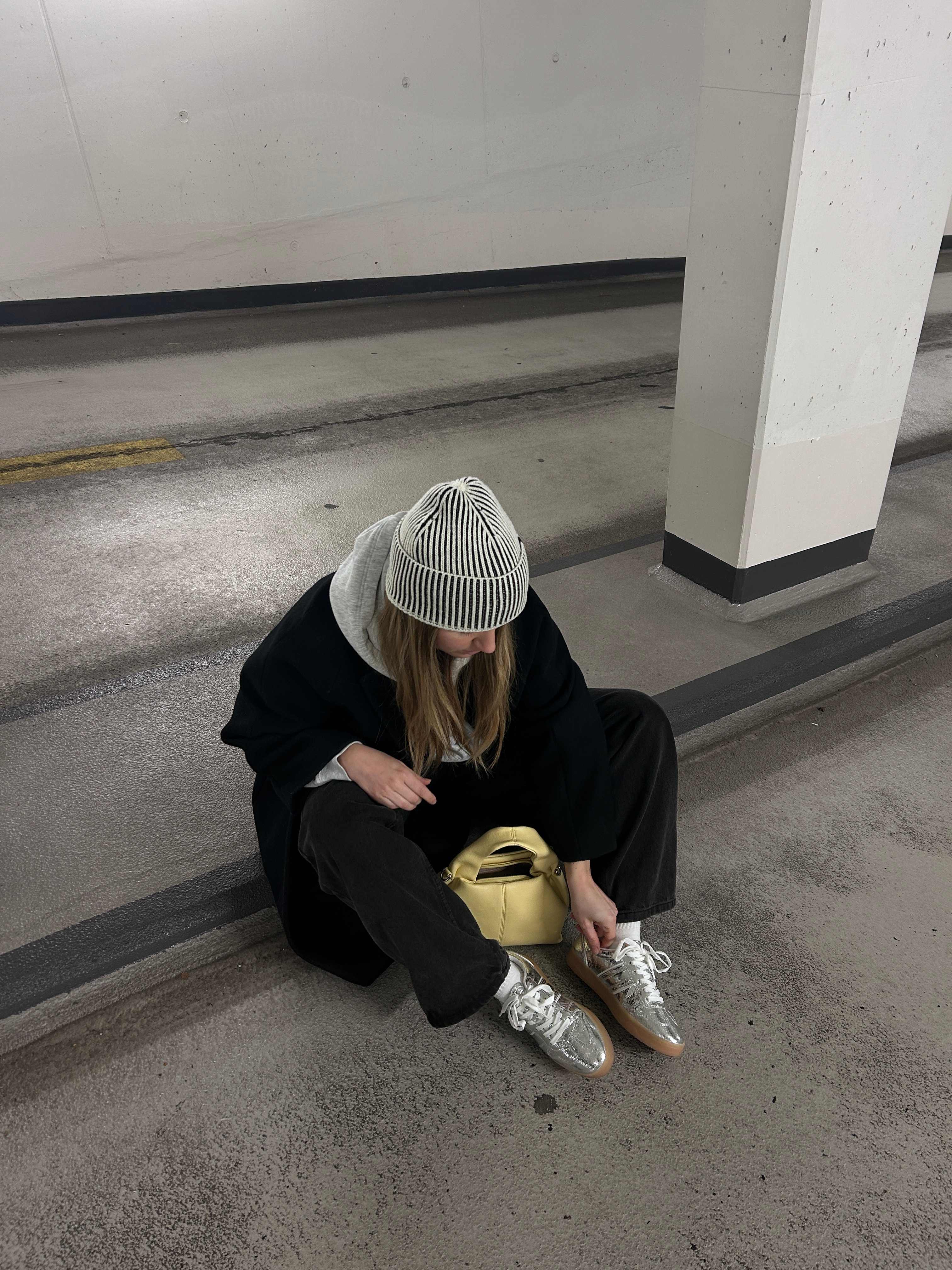 Frau sitzt auf Parkhaus-Rampe, trägt Sneaker in Metallic zu schwarzem Mantel