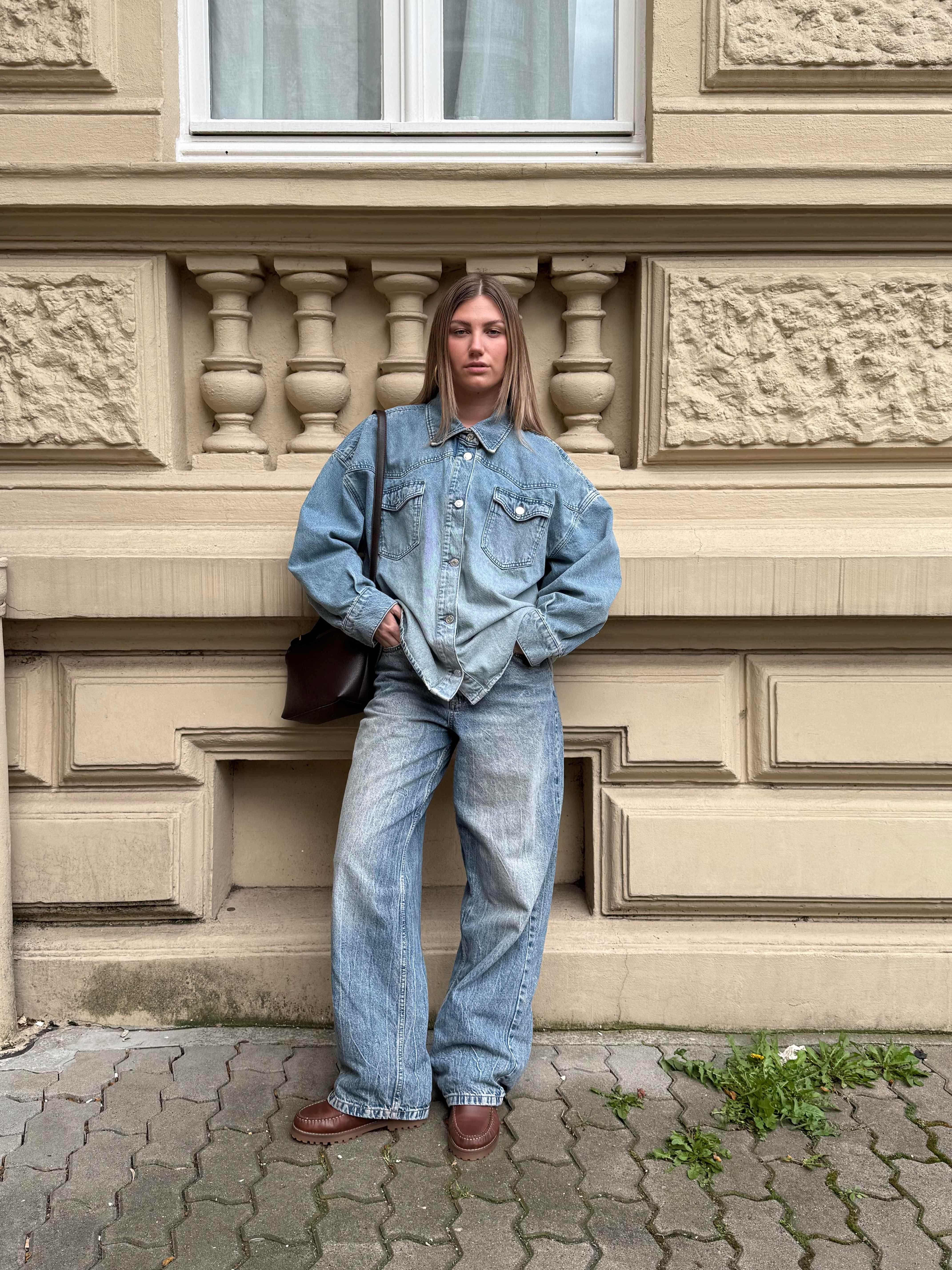 Frau in Denim-Look-Outfit, mit braunen Bootsschuhen, steht lässig vor Wand