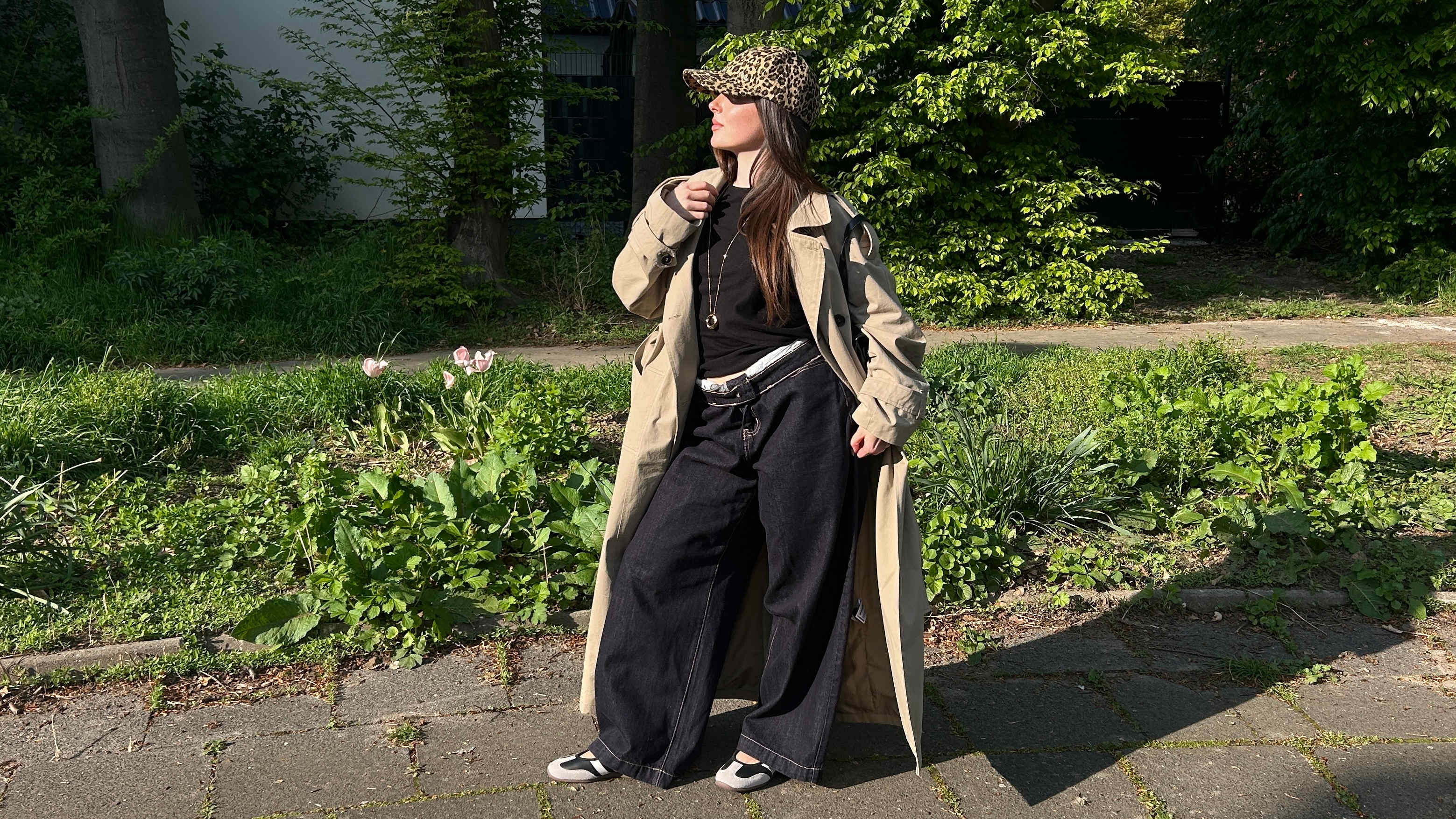 Frau in braunem Trenchcoat, mit schwarzem Jumpsuit und Trend Cap, posiert in Park