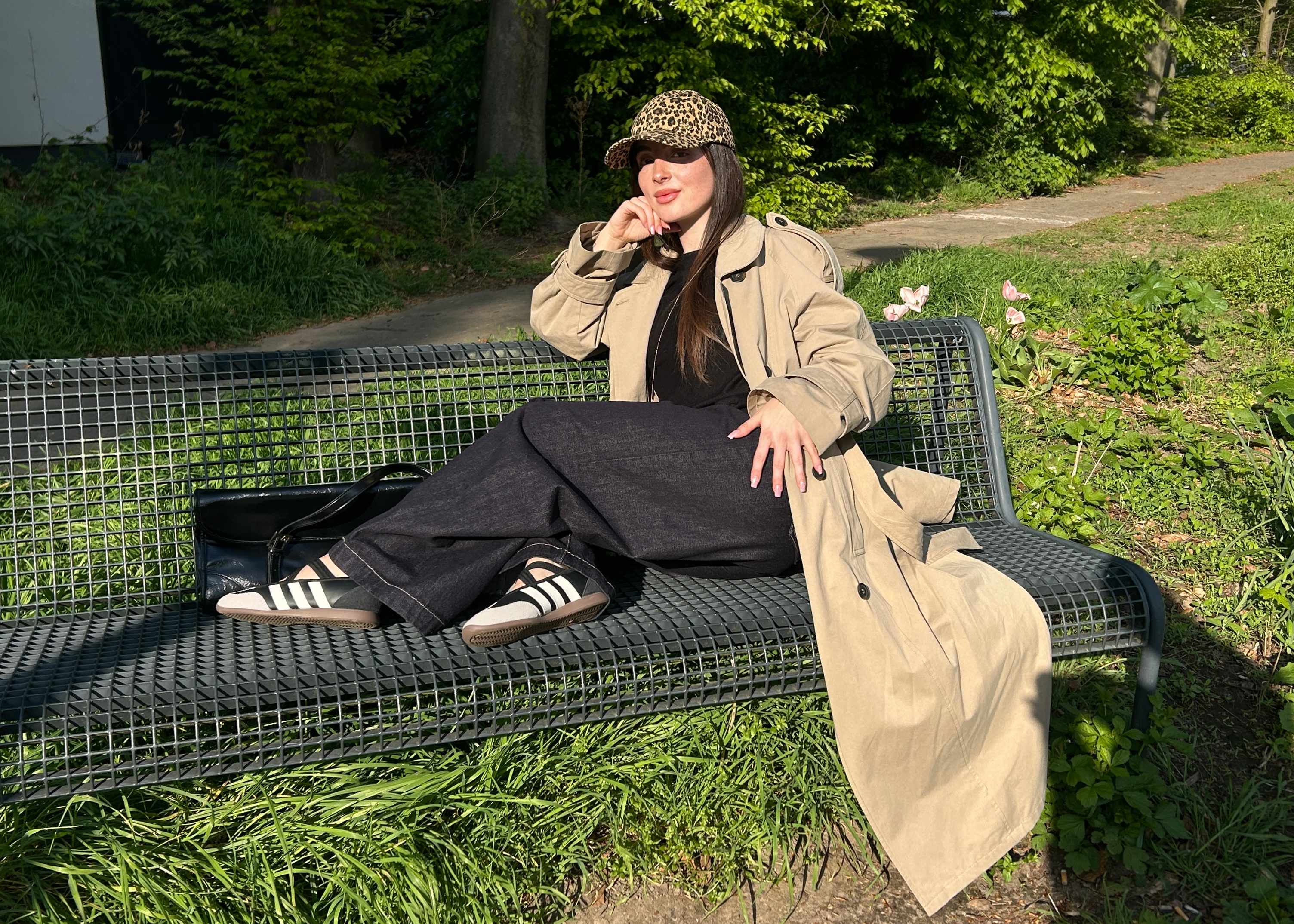 Frau mit braunem Oversize-Coat, trägt schwarze Hose und Sneaker, sitzend auf Parkbank