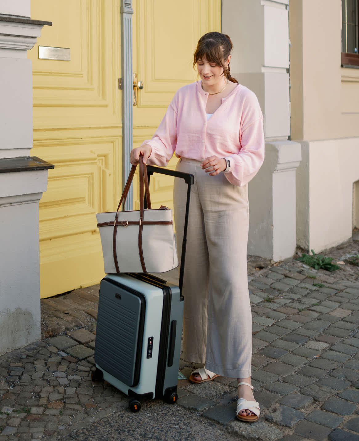 Frau in rosa Flugzeug-Outfit mit grauer Hose neben Rollkoffer