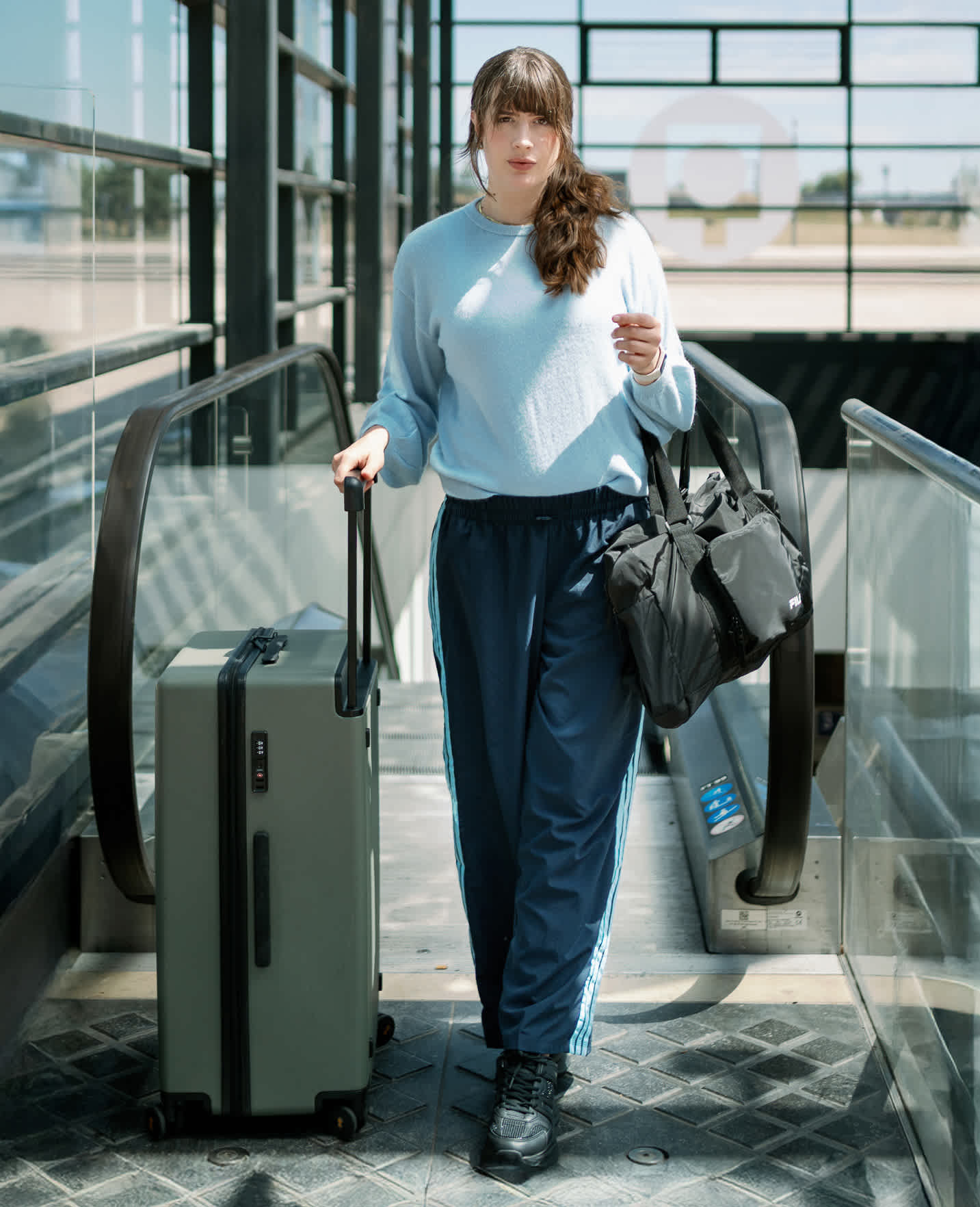 Frau in blauem Sweatshirt, mit Rollkoffer am Arm, am Ende einer Flughafen-Rolltreppe