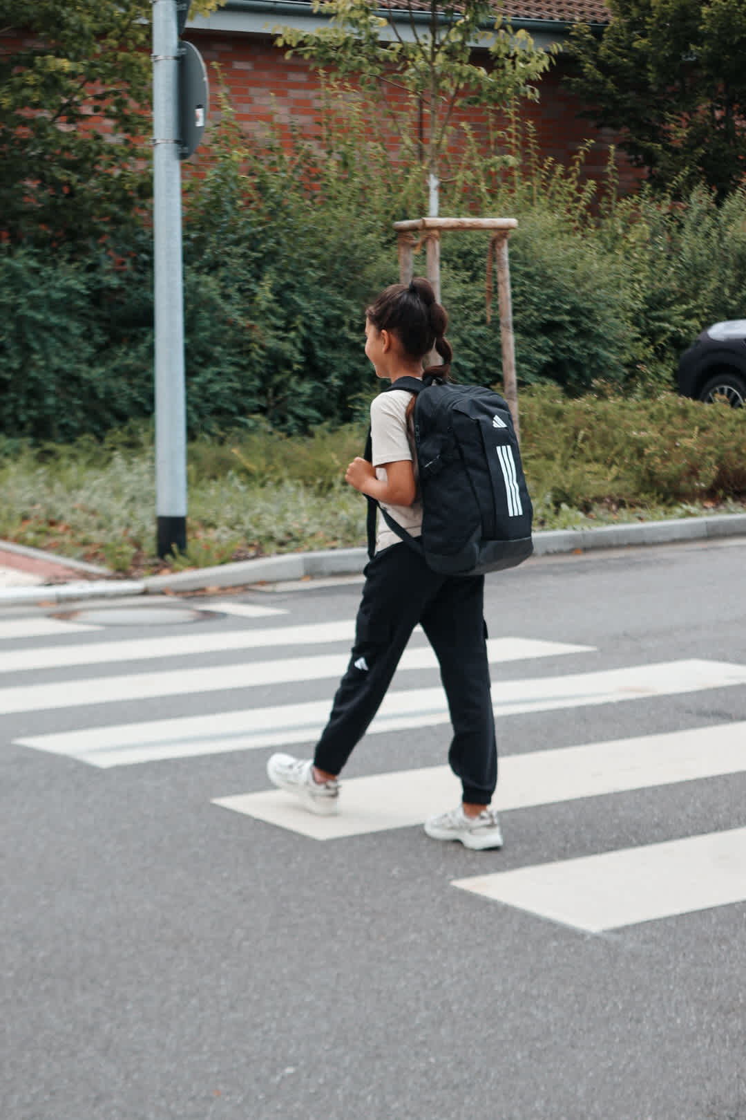 Kind mit Adidas-Rucksack und weißen Sneakern überquert Zebrastreifen auf dem Schulweg