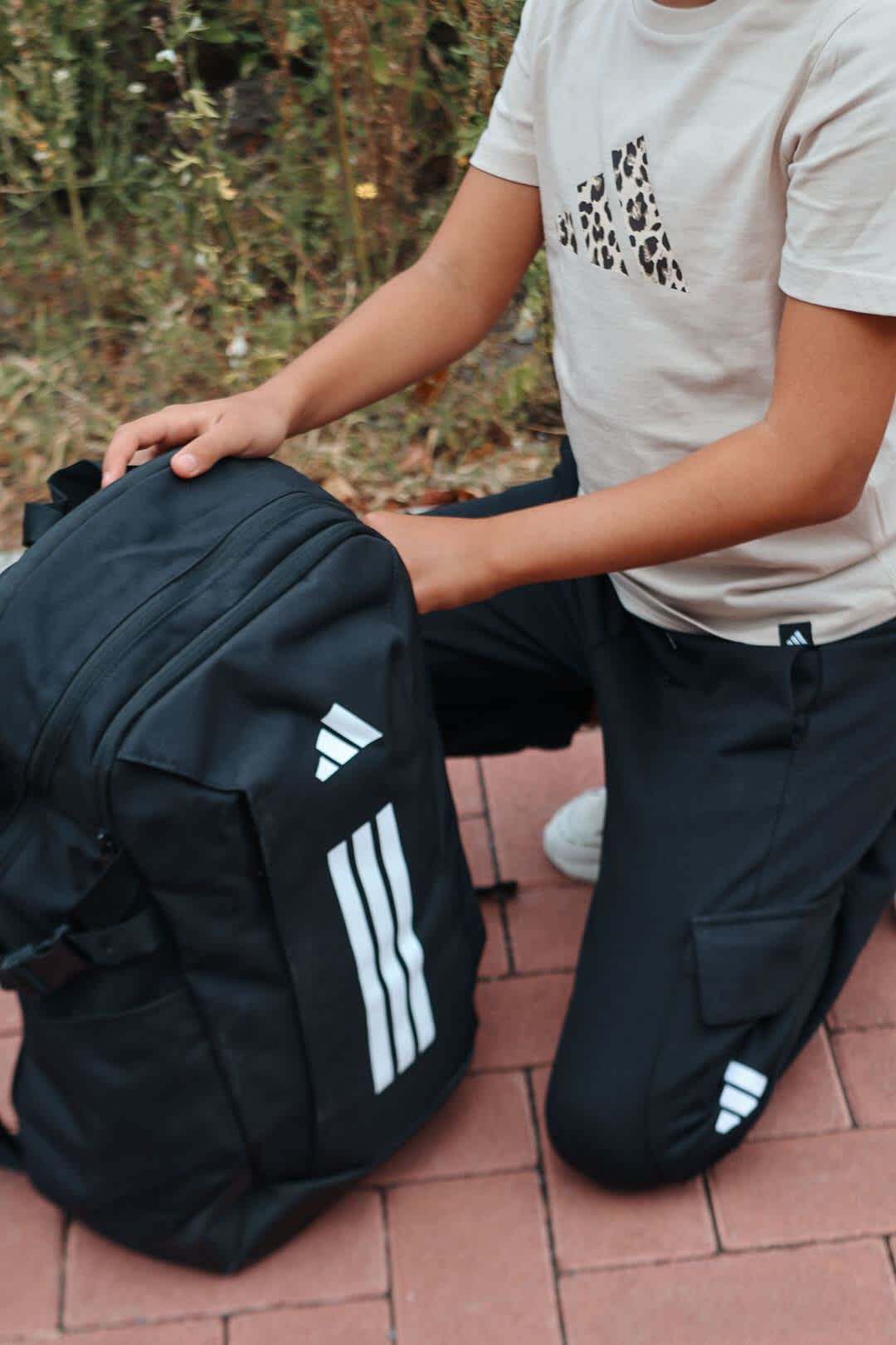 Kind in weißem Adidas-Shirt kniet neben schwarzem Adidas-Rucksack