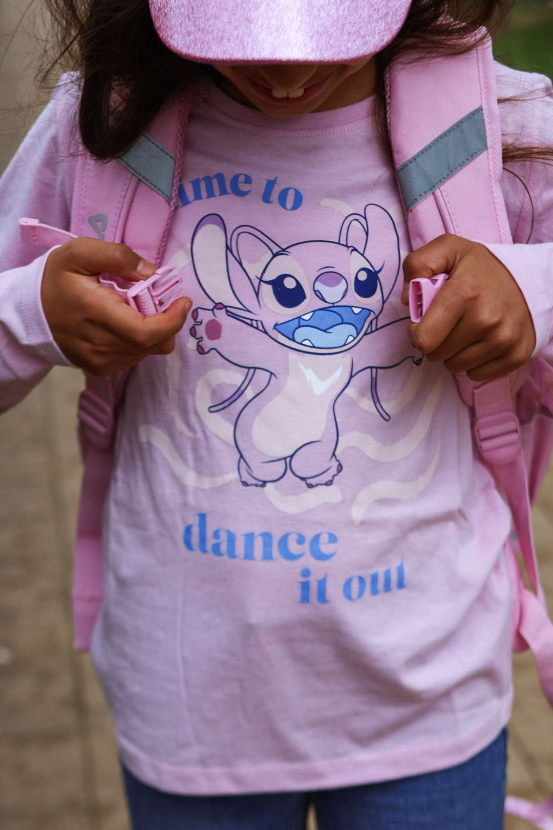 Kind mit rosa Kappe schließt Rucksackgurte, trägt pinkes T-Shirt mit Comic-Motiv und Schriftzug