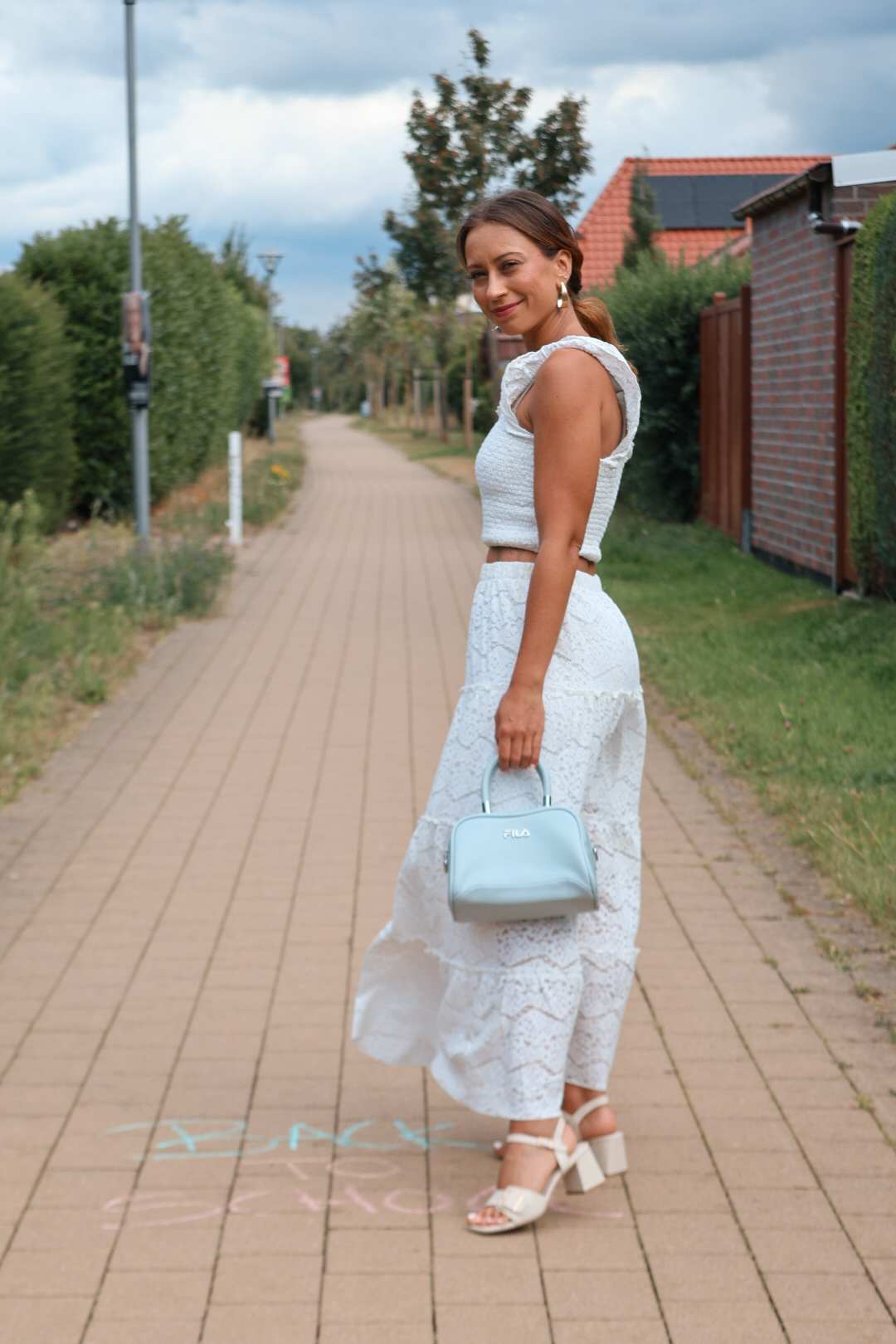 Junge Frau trägt weißes Spitzen-Set mit Fila-Handtasche und weißen Blockabsatz-Sandaletten als Back to School-Outfit