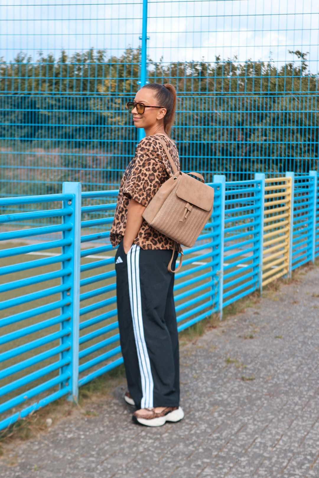 Frau trägt Leo-Print-Shirt mit Adidas-Trainingshose und beigem Rucksack vor blauem Zaun