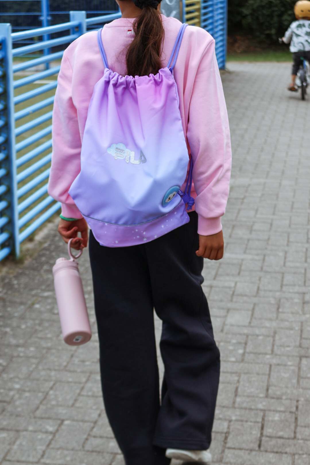 Kind mit lila Fila-Turnbeutel und rosa Trinkflasche läuft über gepflasterten Schulhof