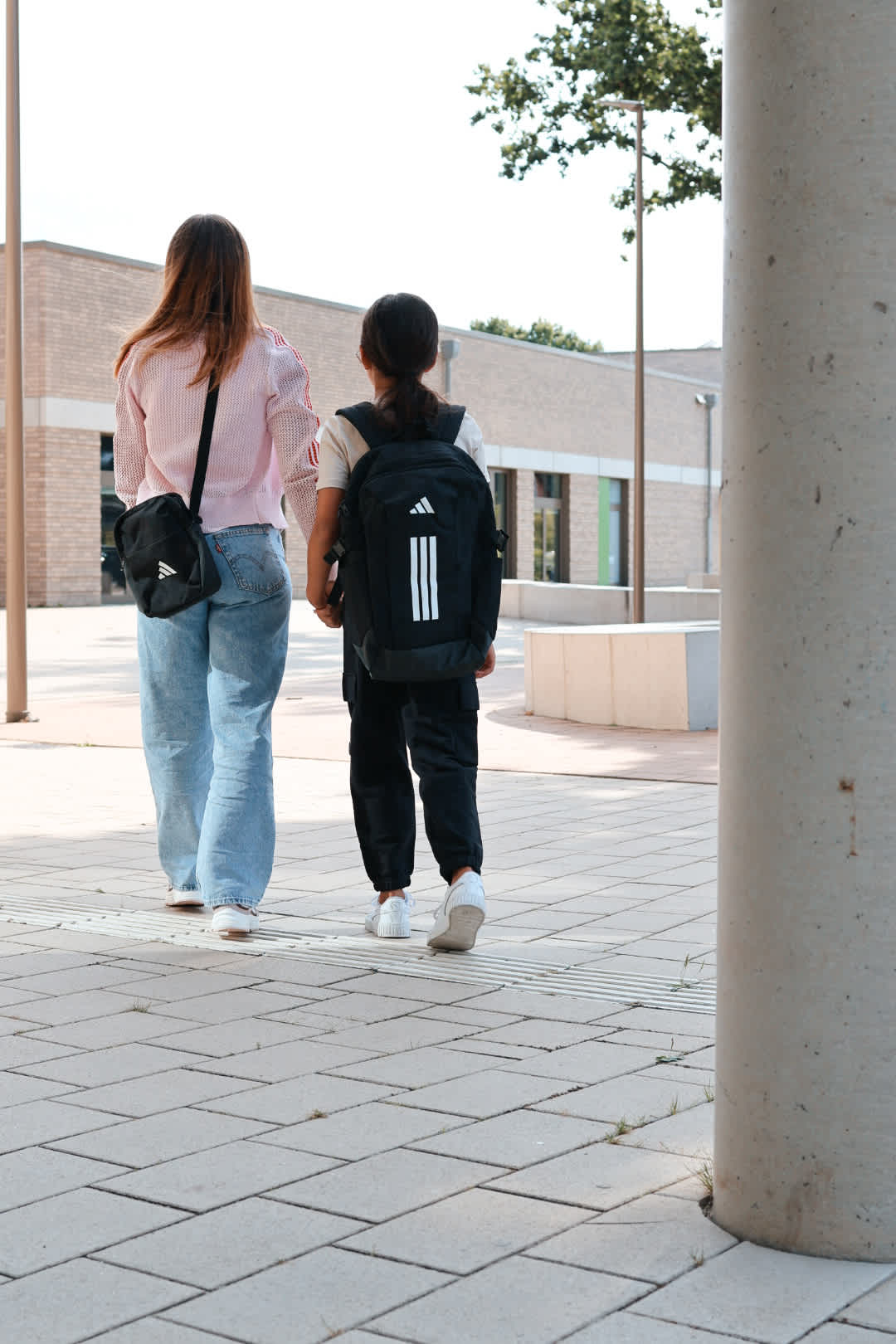 Erwachsene und Kind mit Adidas-Rucksack und Back to School-Outfits laufen händchenhaltend zur Schule