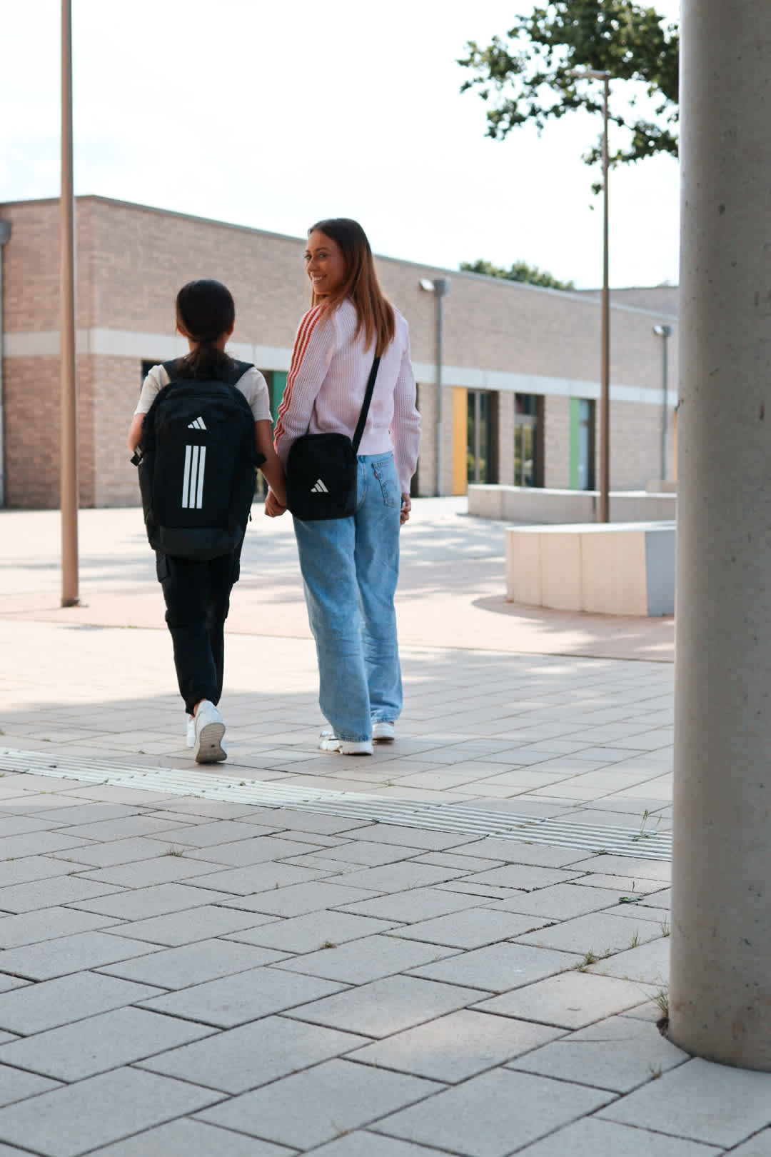 Lächelnde Erwachsene und Kind mit Adidas-Rucksack laufen Hand in Hand zur Schule