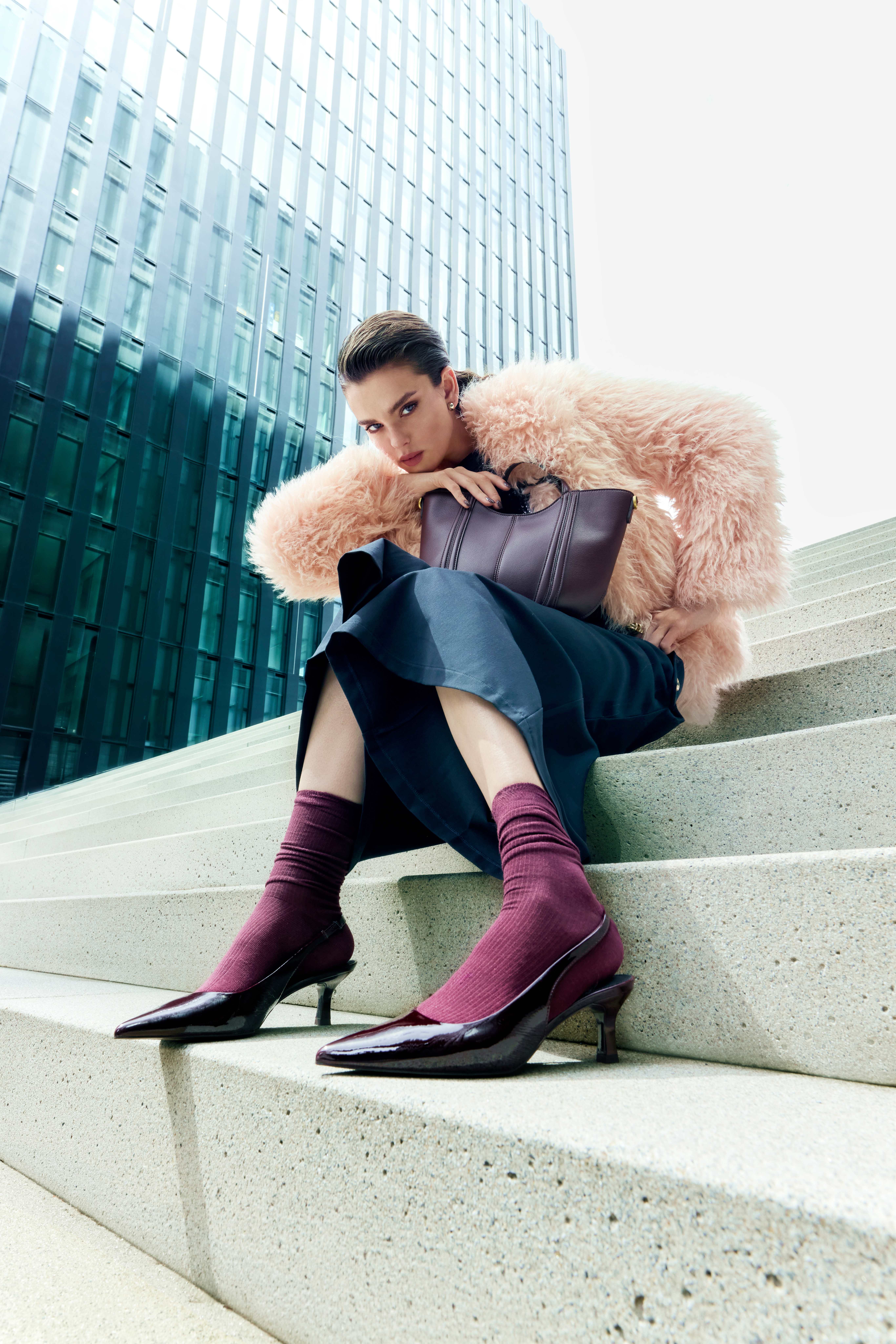 Rosafarbener Oversize-Mantel, dunkelblaues Midikleid und burgunderfarbene Söckchen mit glänzenden Pumps auf Betontreppen