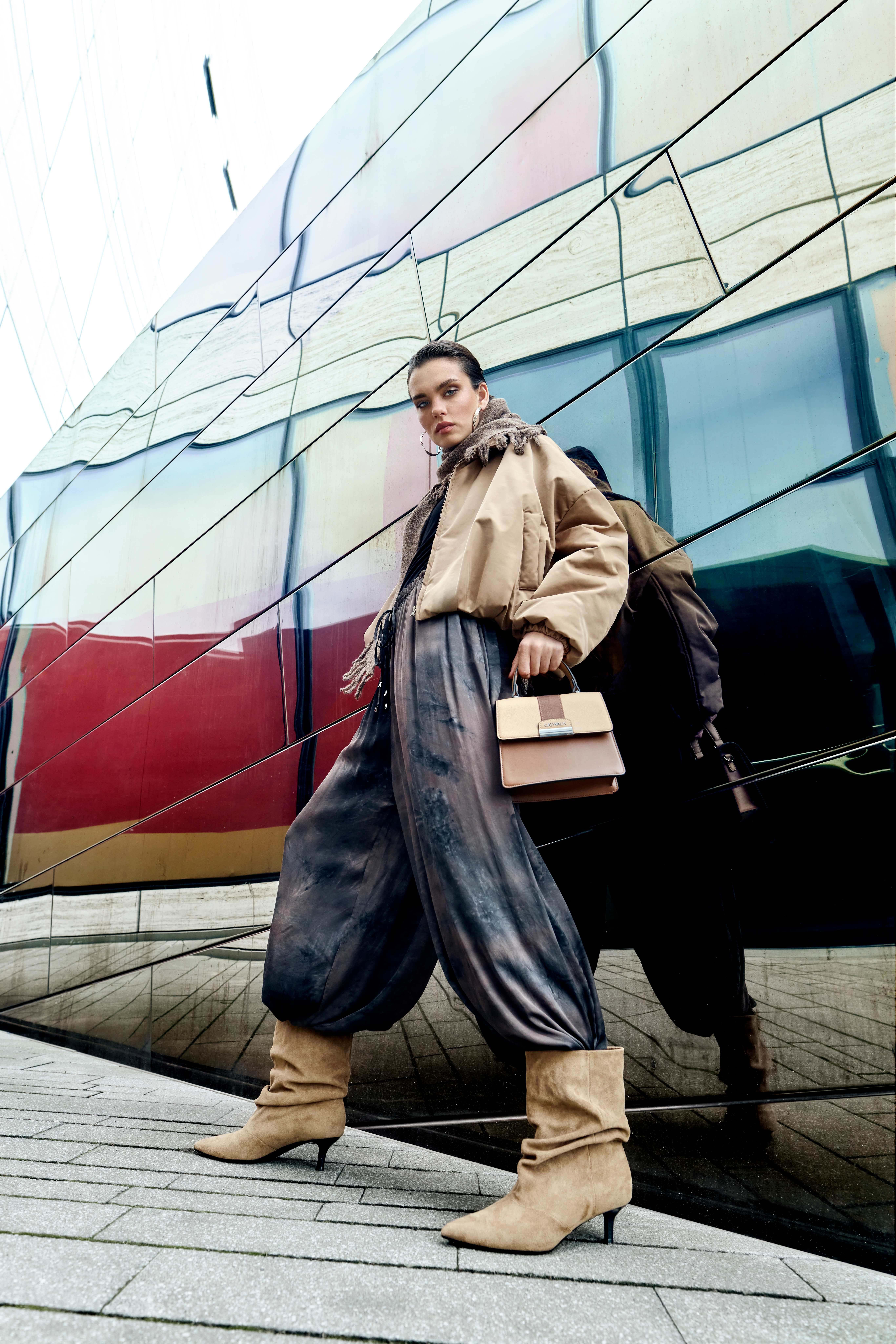 Beiger Oversize-Look mit glänzender Hose und kniehohen Stiefeln vor moderner Architektur